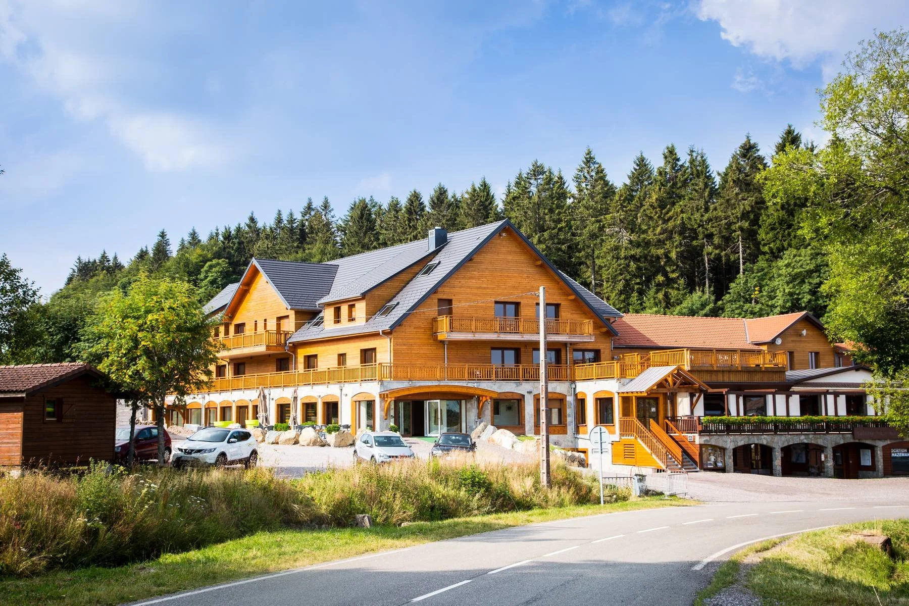 Grand bâtiment en bois avec balcon, situé dans un environnement naturel avec arbres et voiture à l'avant