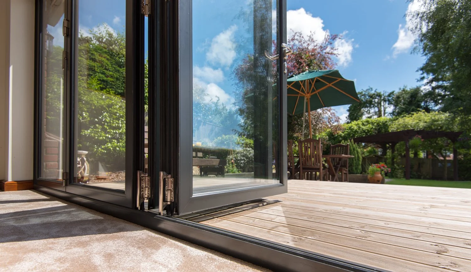Open glass door leading to a wooden deck with outdoor furniture, umbrella, and garden with trees and blue sky