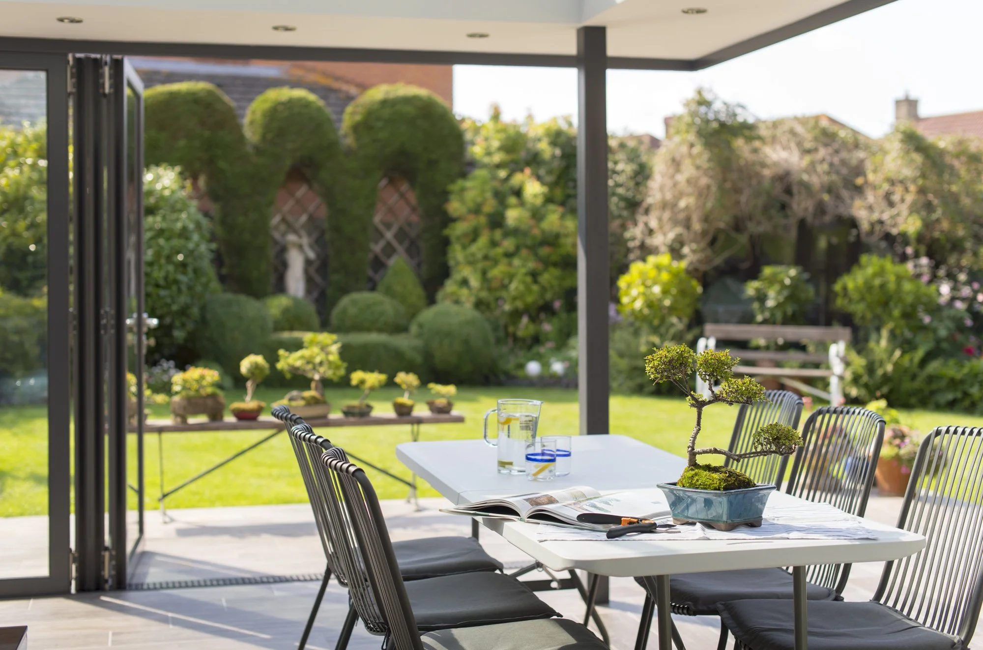 A backyard patio with a white table, black chairs, a bonsai tree, and glasses of water. The patio opens to a lush green garden with trimmed bushes, trees, and a lawn.
