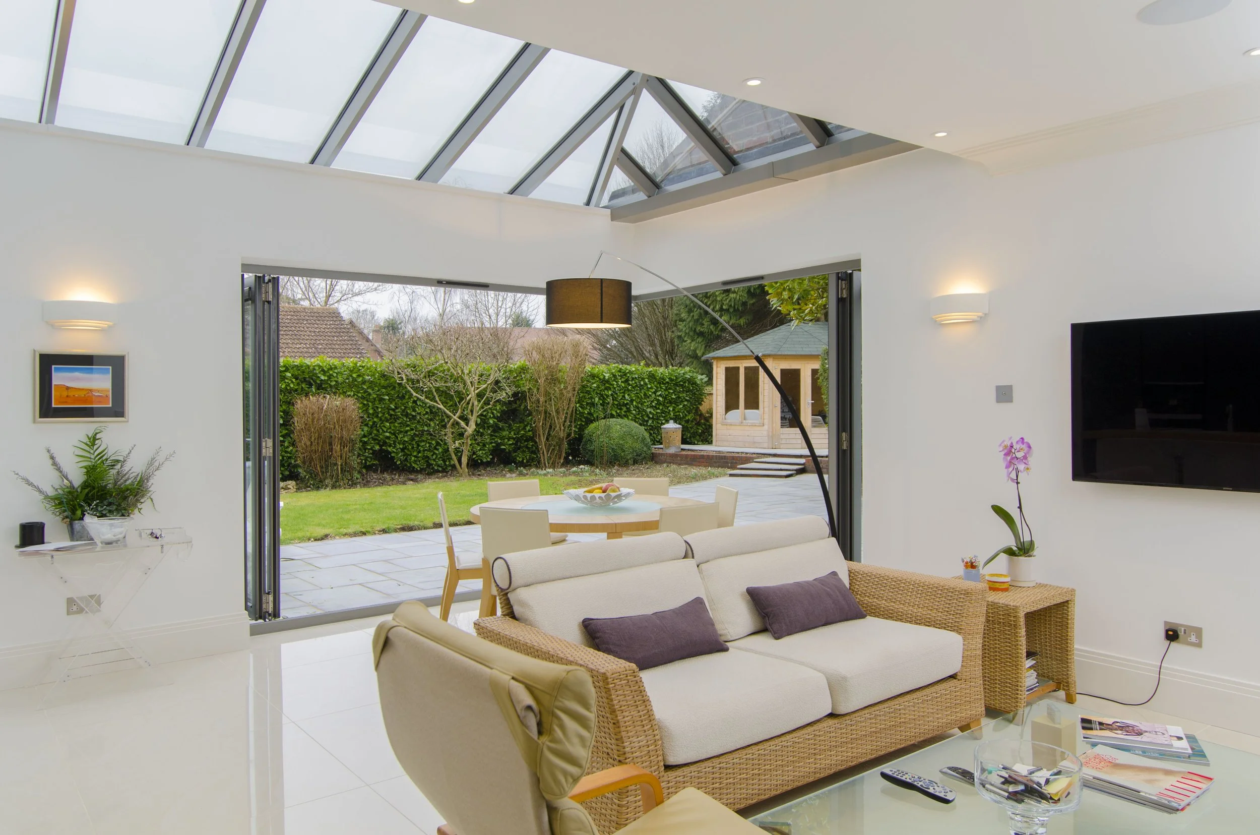 Living room with large glass doors opening to a backyard with a garden and shed, beige sofa with purple cushions, glass coffee table with magazines, TV on the wall, and a skylight ceiling.