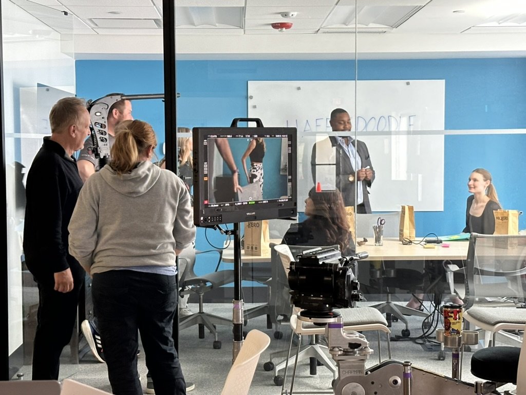 A film or television scene being shot in a conference room with a glass wall. A man in a suit is speaking at a whiteboard, while three women are seated at a table with gift bags. Crew members and a camera operator are filming the scene, with equipment set up in front.