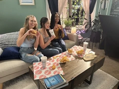 Three women sitting on a sofa enjoying fast food, including fried chicken and drinks, in a cozy living room set
