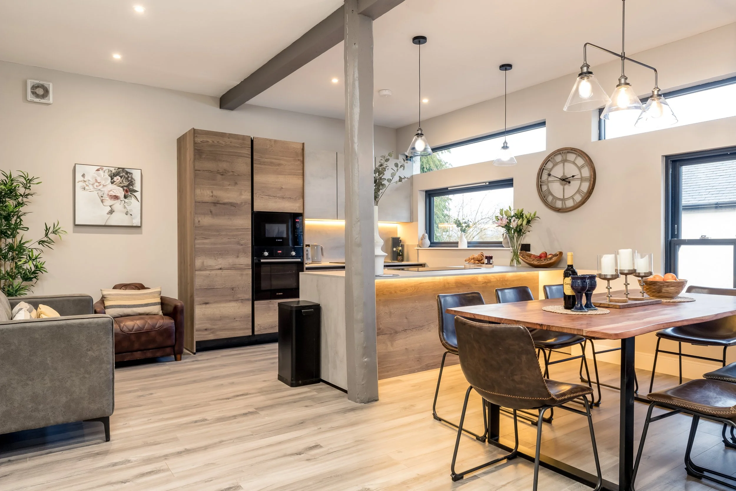 Open-concept living and dining area with large windows, modern decor, a clock on the wall, and a kitchen with a wooden counter and pendant lights.