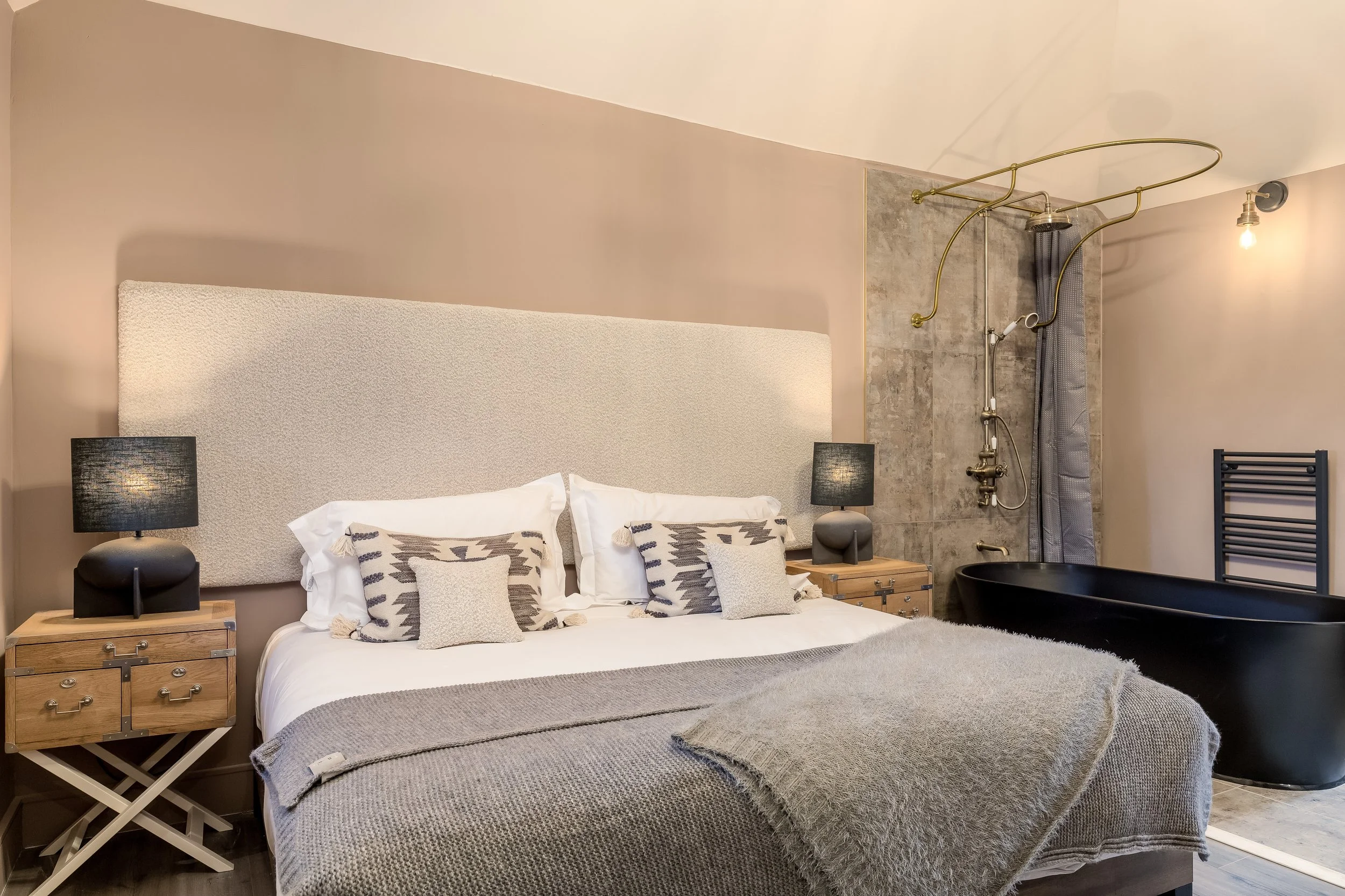 A bedroom with a large bed, nightstands with black lamps on each side, decorative pillows, a textured headboard, and a bathroom area with a shower, curtain, towel radiator, and black bathtub.