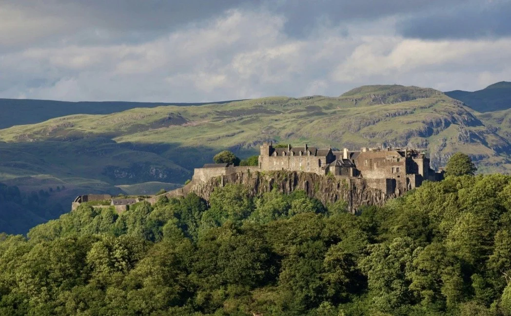 A historic castle on a green hill surrounded by trees with rolling green hills in the background under a partly cloudy sky.