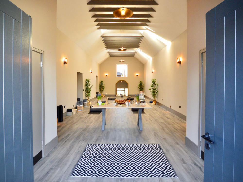 A spacious dining area with a long table set against a high ceiling with wooden beams, illuminated by ceiling lights and wall sconces, decorated with potted plants along the sides, and a patterned rug at the entrance.