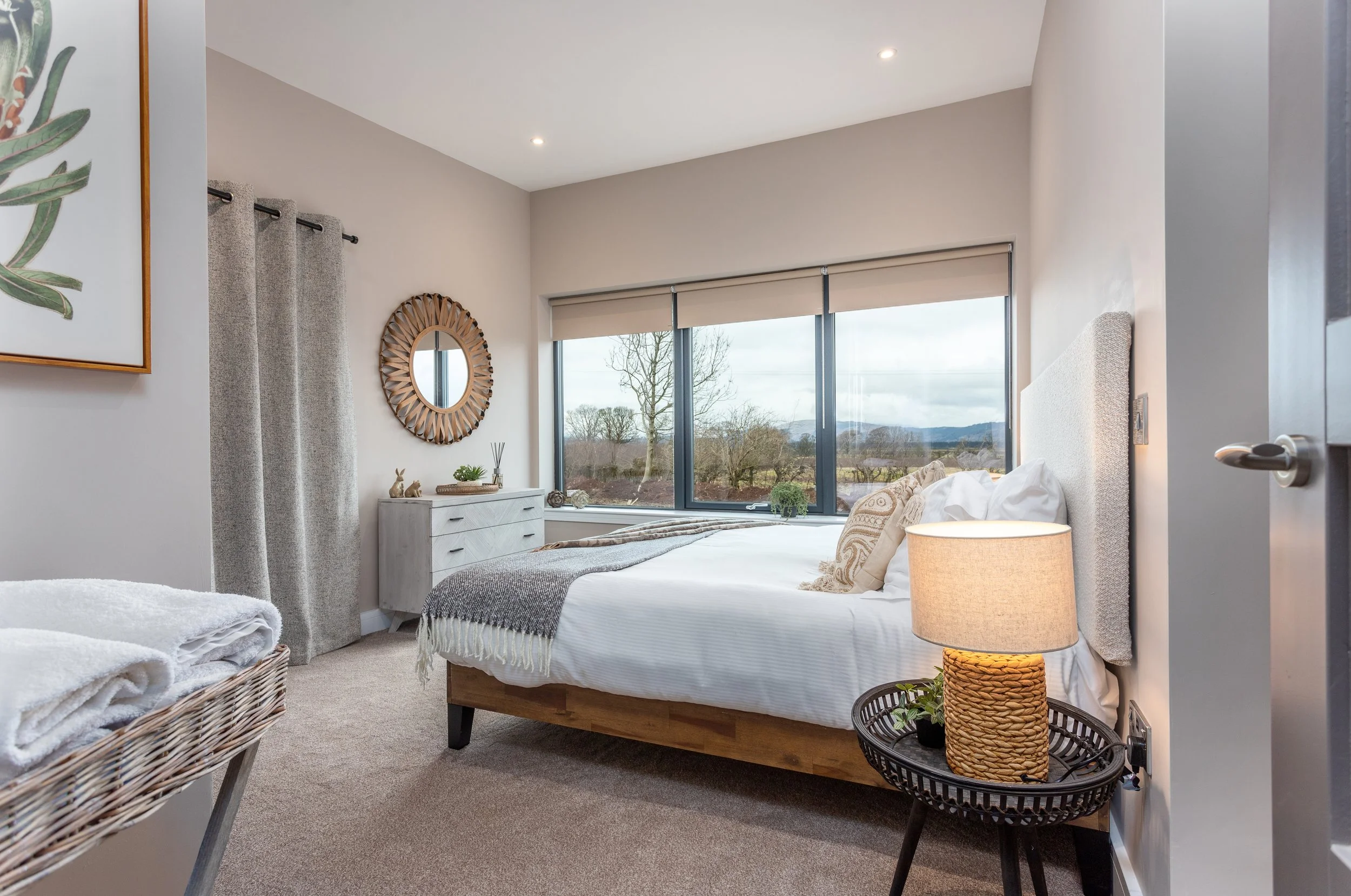A bedroom with a large window revealing a scenic landscape outside. The bed has white linens, beige pillows, and a gray throw blanket. A wicker basket with towels and a woven table with a lamp and plants are near the bed. The wall features a round mirror and a framed botanical print, with beige curtains hanging beside the window.
