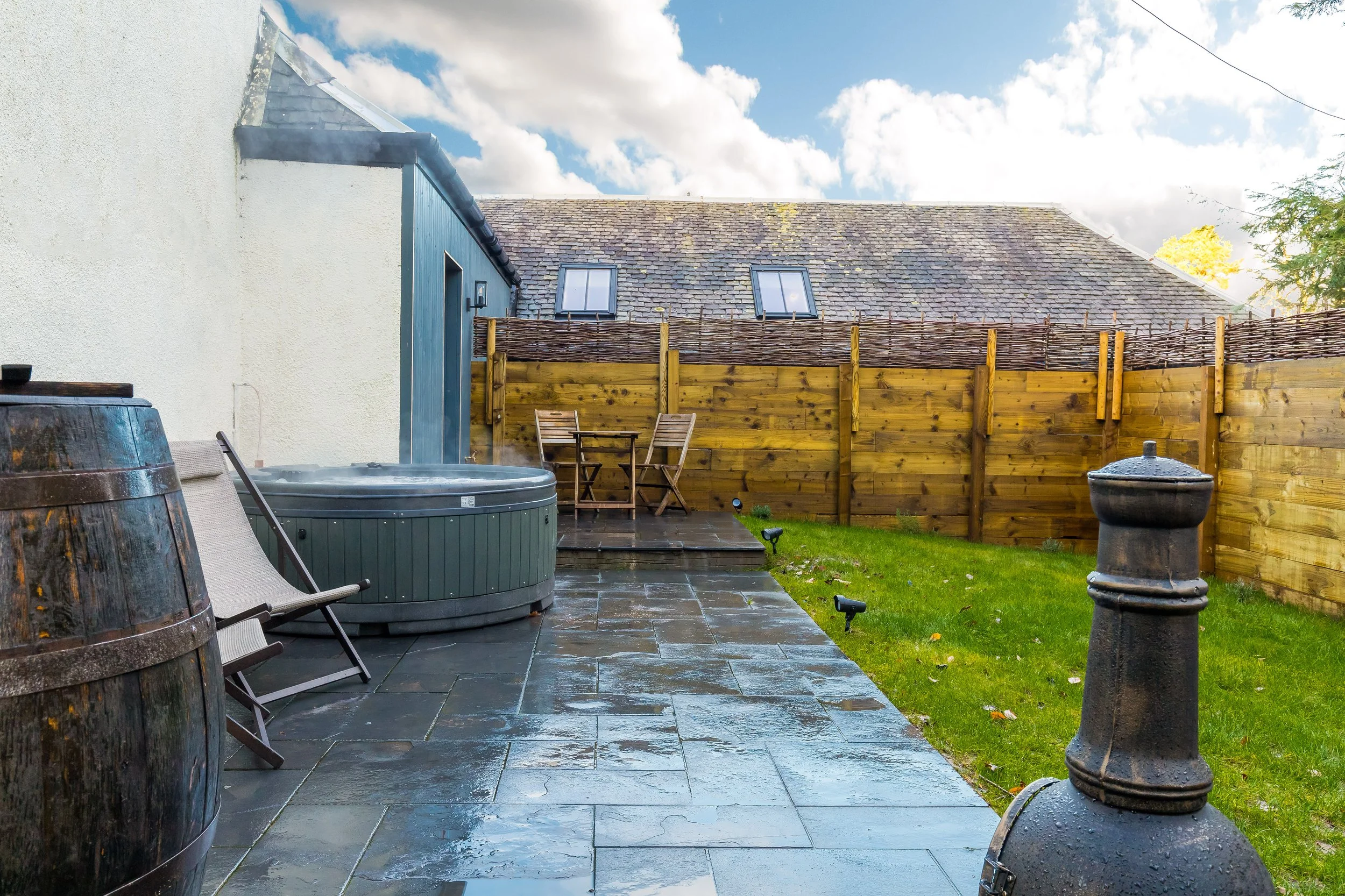 A backyard patio with a hot tub, two chairs, a wooden fence, and a house with a sloped roof and two windows. The sky is partly cloudy and the ground appears wet.