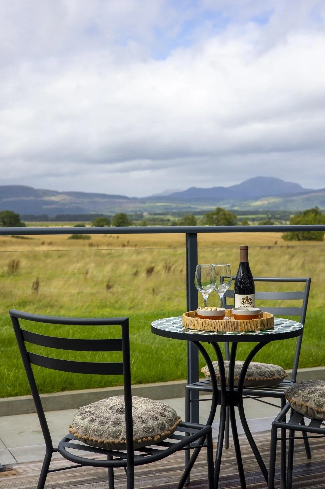Outdoor patio with a small round table, two chairs with patterned cushions, a bottle of wine, two glasses, and small bowls, overlooking a green field and distant mountains under cloudy sky.