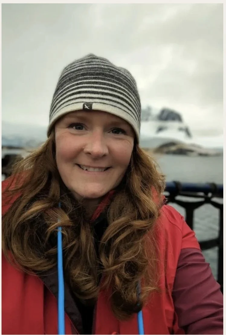 A woman with long wavy hair wearing a striped beanie hat and a red jacket, smiling near a body of water with a boat and cloudy sky in the background.