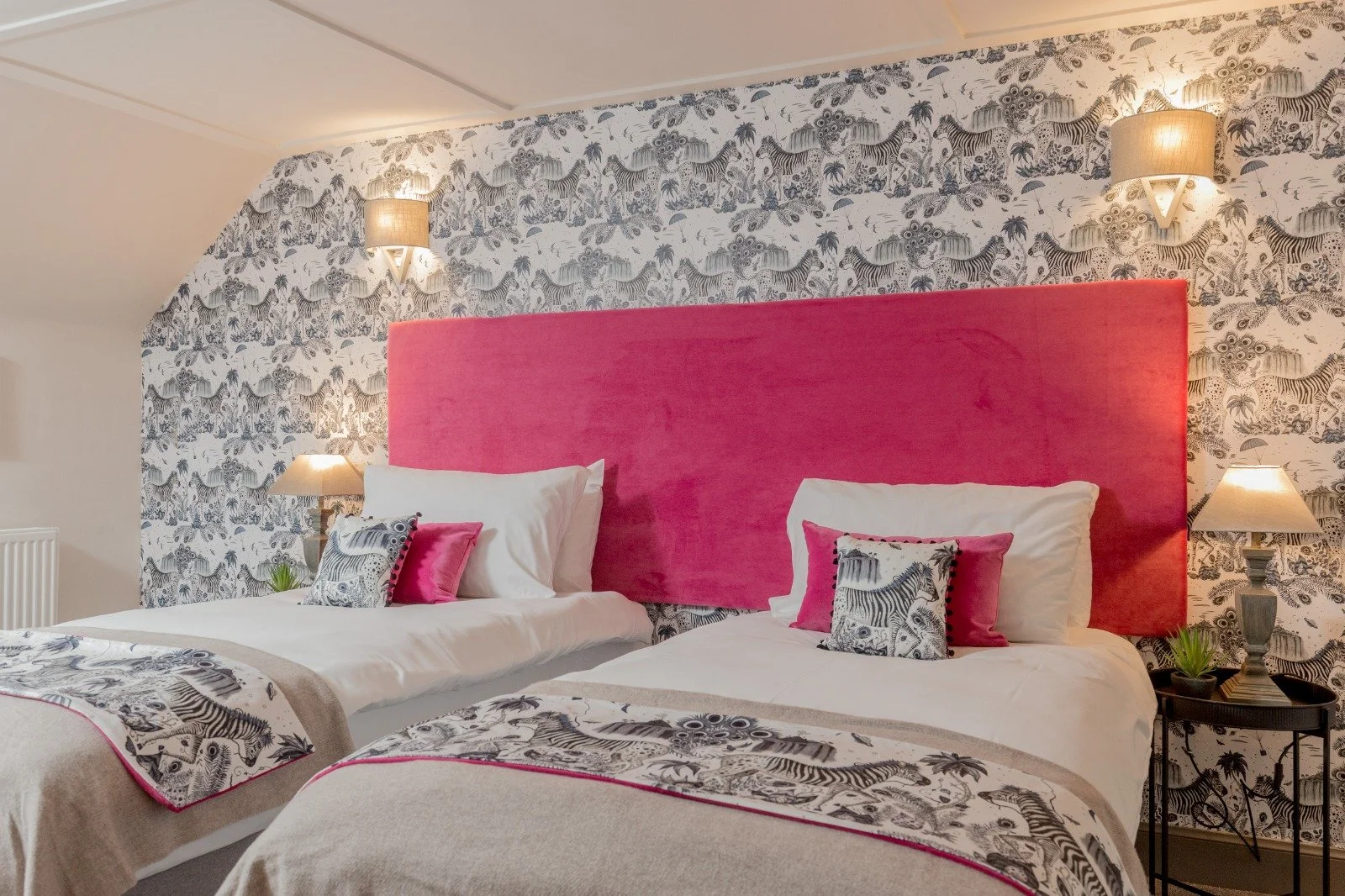Two beds with pink upholstered headboards, white bedding, zebra and palm tree patterned pillows, and matching bed runners. The wall behind has black and white zebra and animal print wallpaper, with two wall-mounted lamps and two bedside tables with lamps and small plants.