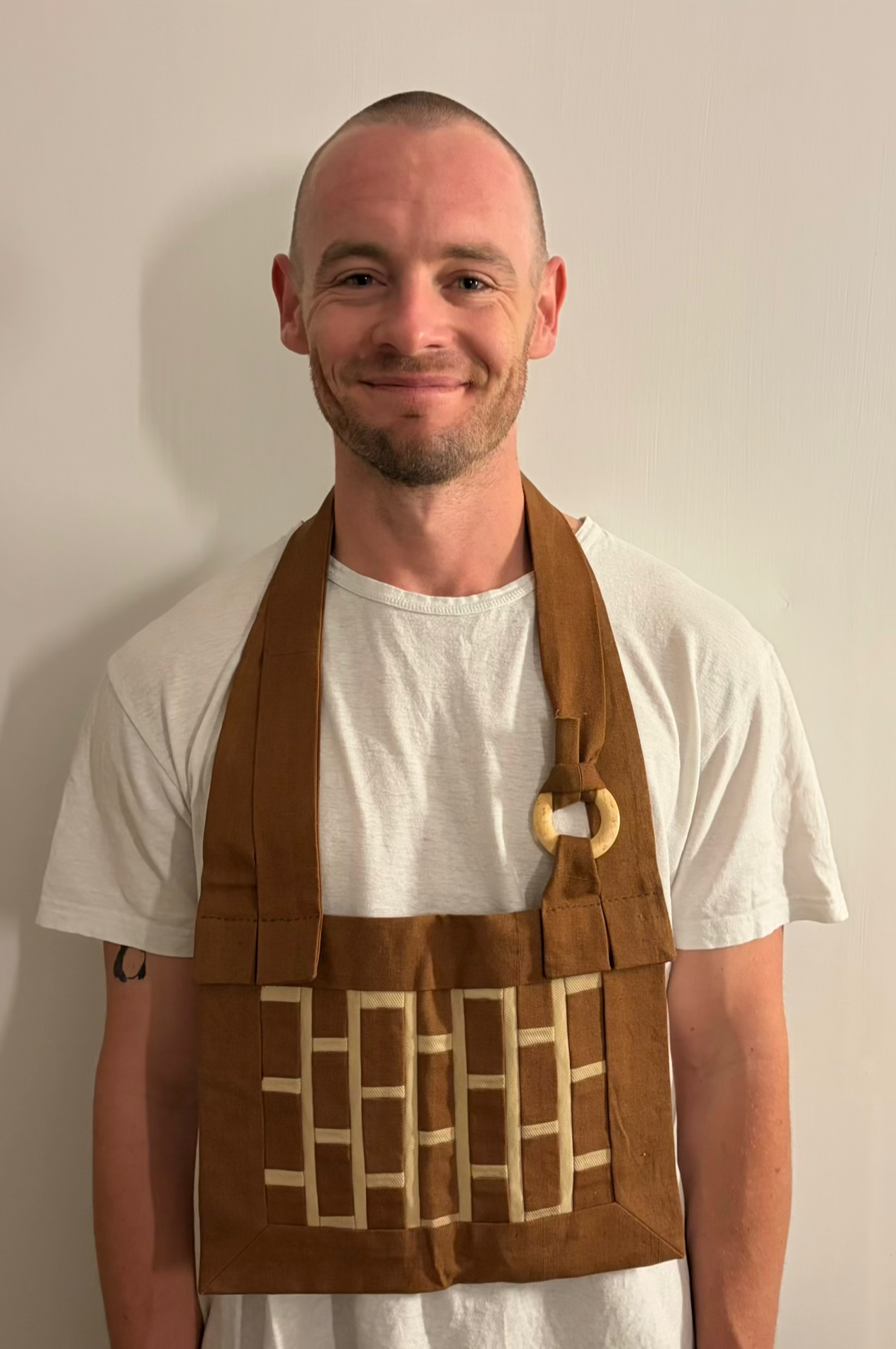 A man with a shaved head and a beard smiling while wearing a beige t-shirt and a brown apron with a checkered pattern.