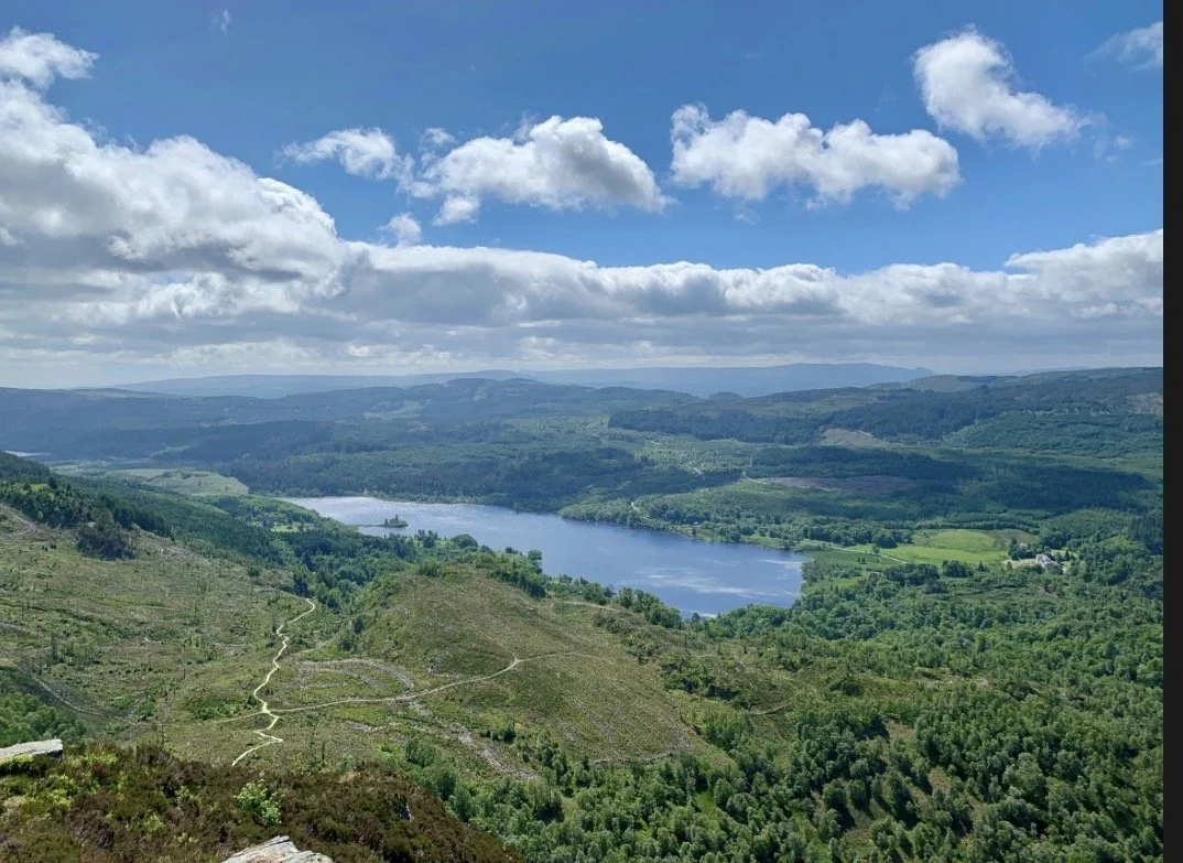 Scenic view of a large lake surrounded by lush green hills and forests under a blue sky with scattered clouds.