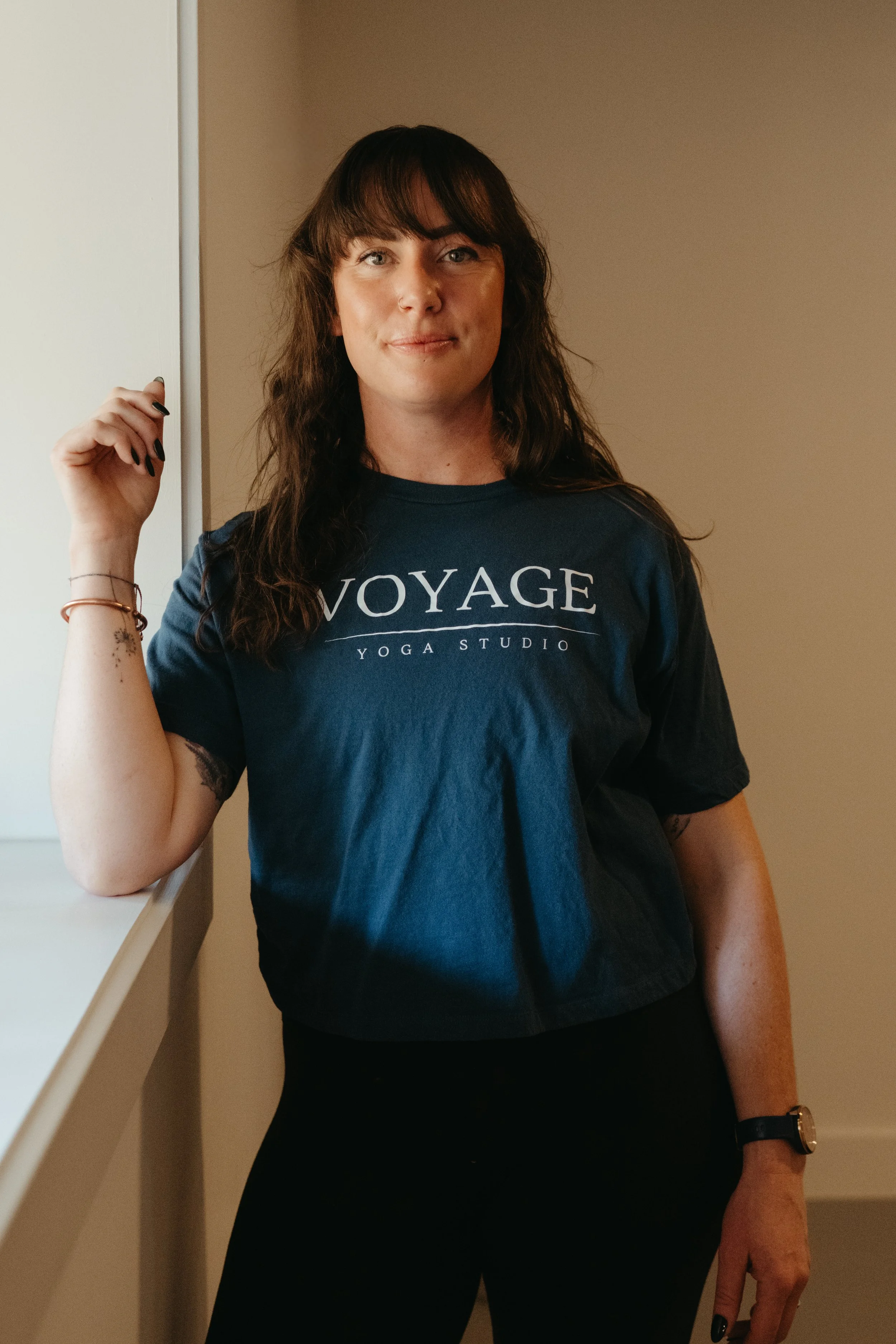 A woman with brown hair wearing a navy blue T-shirt with white text that says "VOYAGE YOGA STUDIO," standing next to a wall with one arm resting on a ledge and the other hanging down. She has tattoos on her forearm and wears a bracelet and a watch.