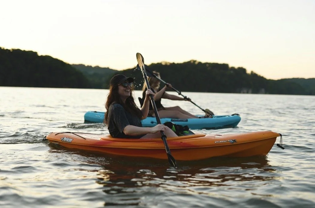 Two women kayaking on a calm lake during sunset with tree-covered hills in the background.