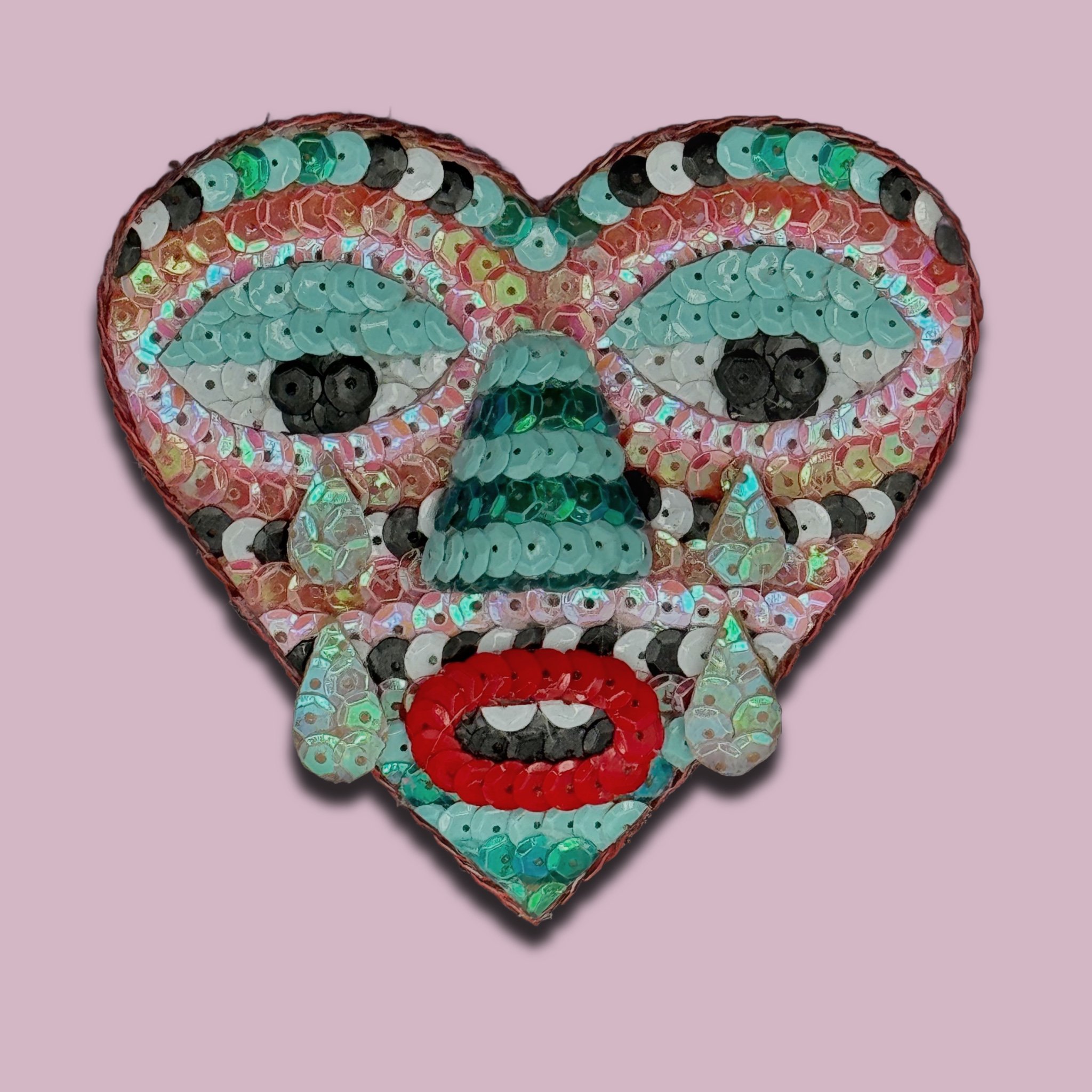 Sequin heart with a clown face design on a pink background.