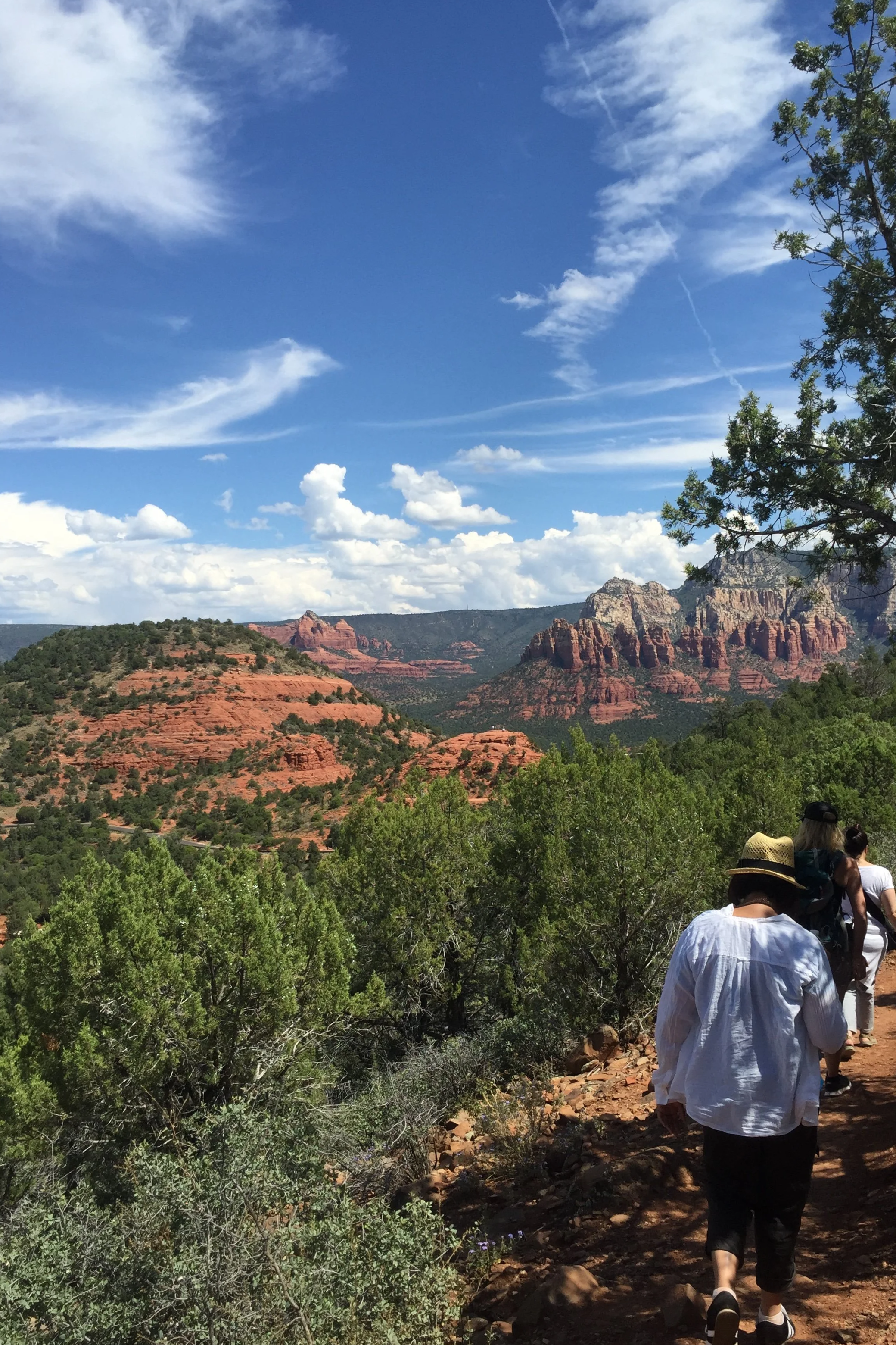 People hiking on a trail through greenery in Sedona, Arizona with red rock formations in the background and a partly cloudy sky above.