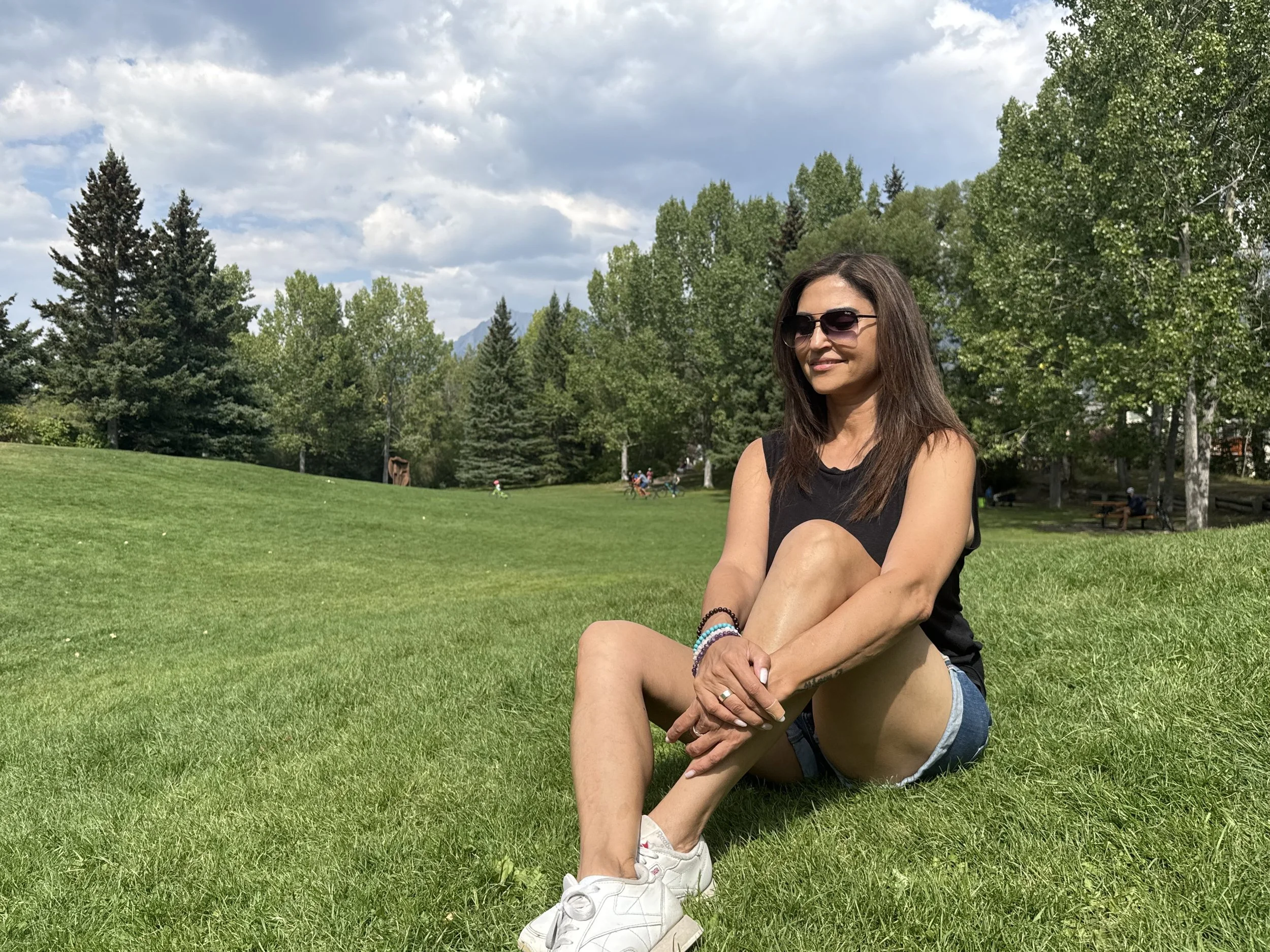 Arzeen Asaria, Spiritual & Mindset Couch sitting on a lush green lawn in a park. There are tall trees and a cloudy sky in the background, with children playing and people sitting on benches.