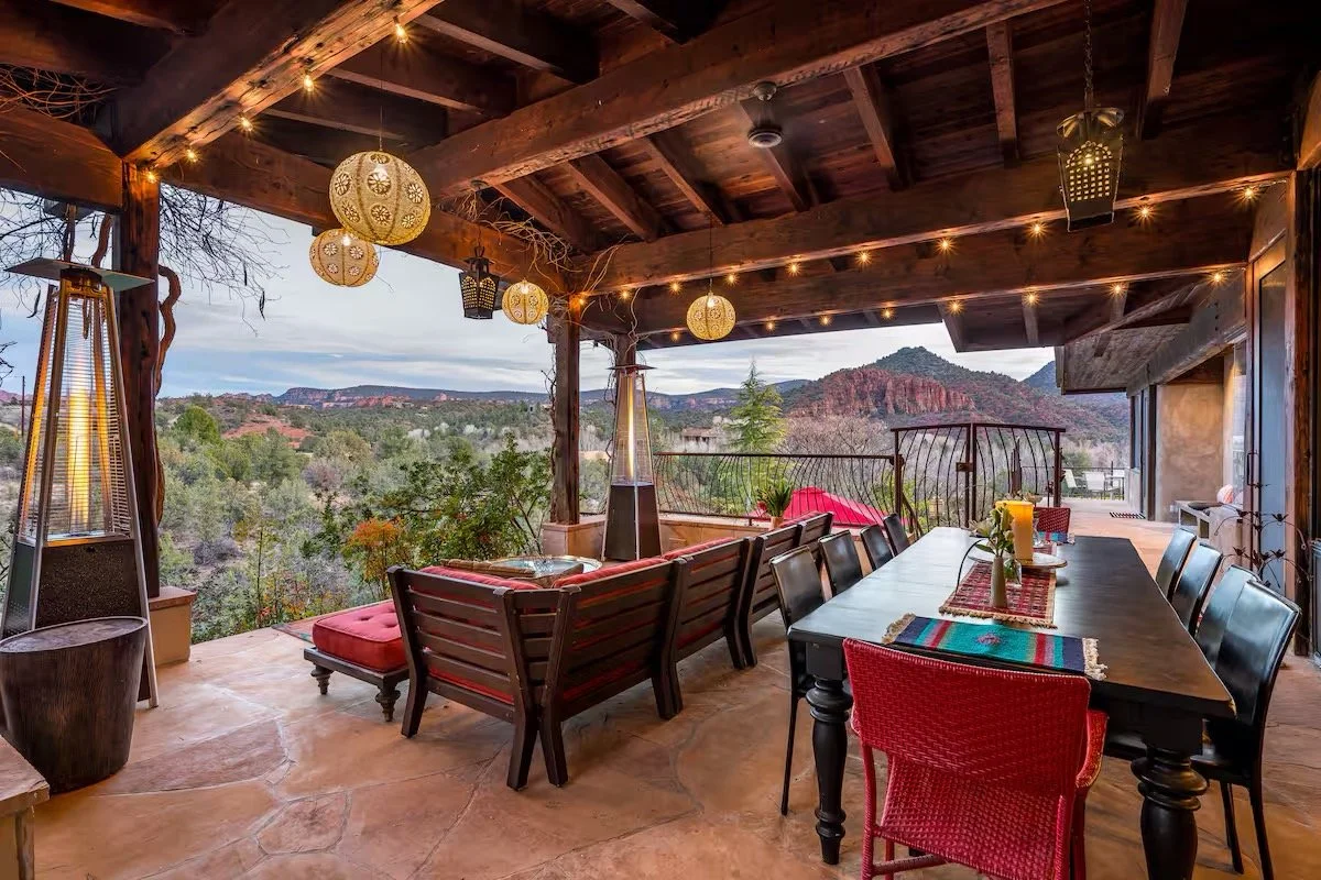 An outdoor patio with a dining table and chairs, decorated with hanging lanterns and string lights, overlooking a scenic mountain landscape during the evening.