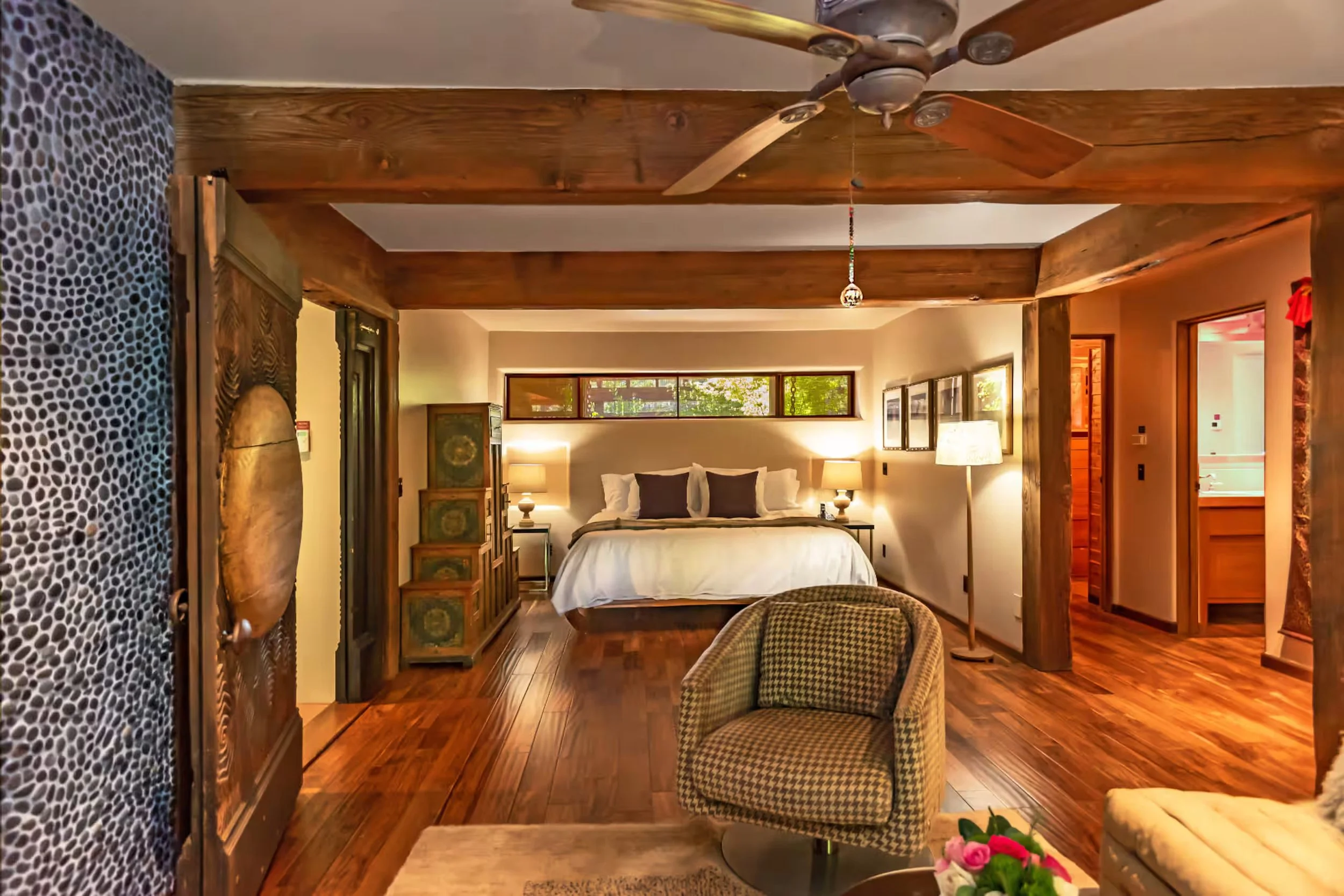 Cozy bedroom with a large bed, wooden accents, and a window with greenery outside. The room has wooden flooring, lamps on bedside tables, and a ceiling fan.