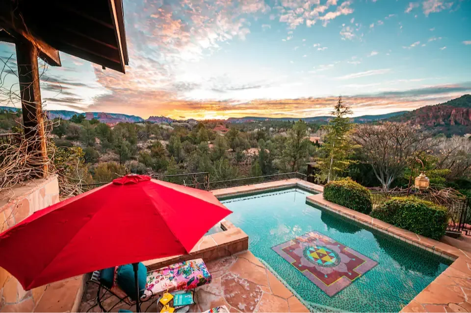 A scenic backyard with a swimming pool, surrounded by trees and hills during sunset. A red umbrella covers a bench with towels and drinks, overlooking the landscape.