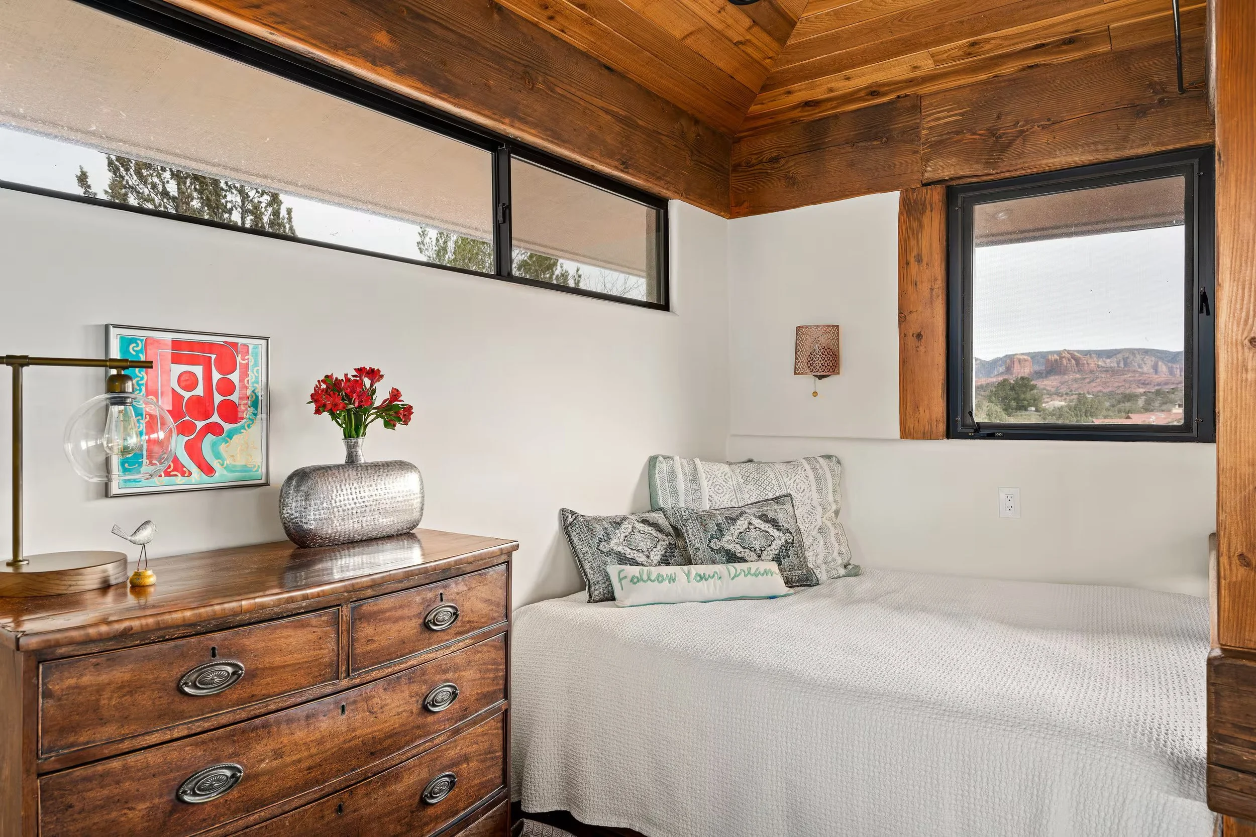 A bedroom with a wooden wall and ceiling, a window showing a landscape with rocks and trees, a dresser with a lamp, a framed colorful artwork, a bouquet of red flowers, and a bed with patterned pillows and a pillow that says 'Follow Your Dreams'.