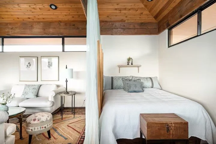 Split view of a cozy living room and bedroom separated by a curtain in a modern home with wooden ceiling and white walls.