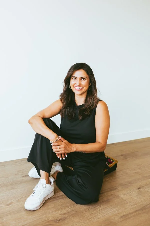 Arzeen Asaria, mindset coach for women, sitting on the floor against a plain white wall, smiling at the camera, dressed in black pants, a sleeveless top, white sneakers, with a colourful box nearby.