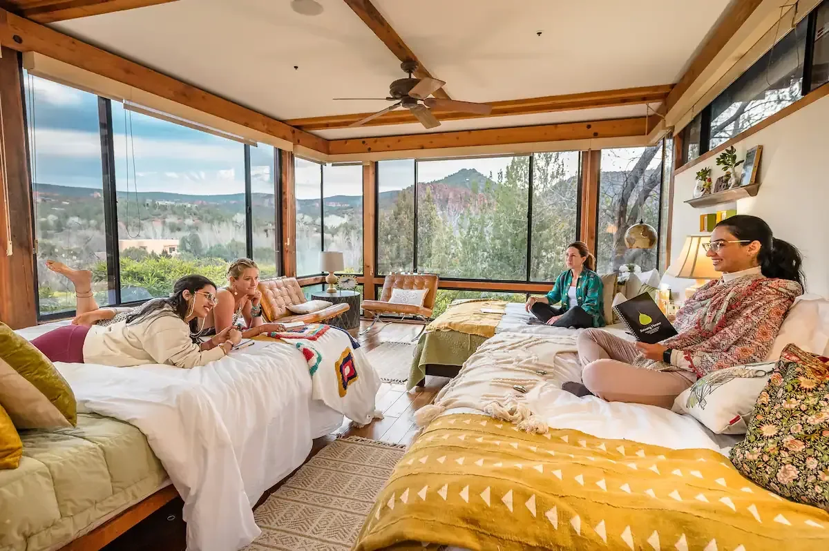 Four women relaxing on beds and couches in a bright room with large windows showing a scenic mountain view, some using laptops and others chatting.