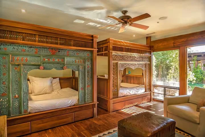 Two wooden canopy beds with decorative carved panels in a sunlit room, with armchair and ottoman.