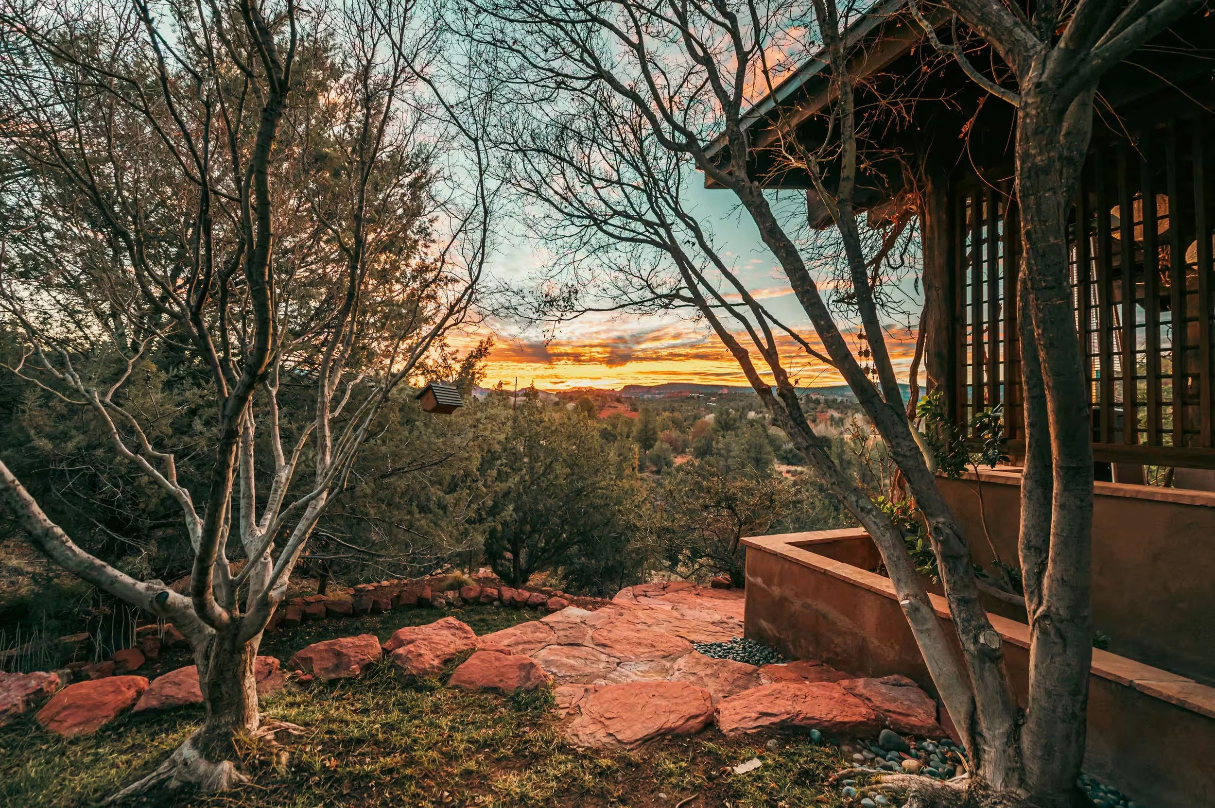 A sunset view from a porch with trees, rocks, and plants, overlooking a distant landscape with hills and trees.