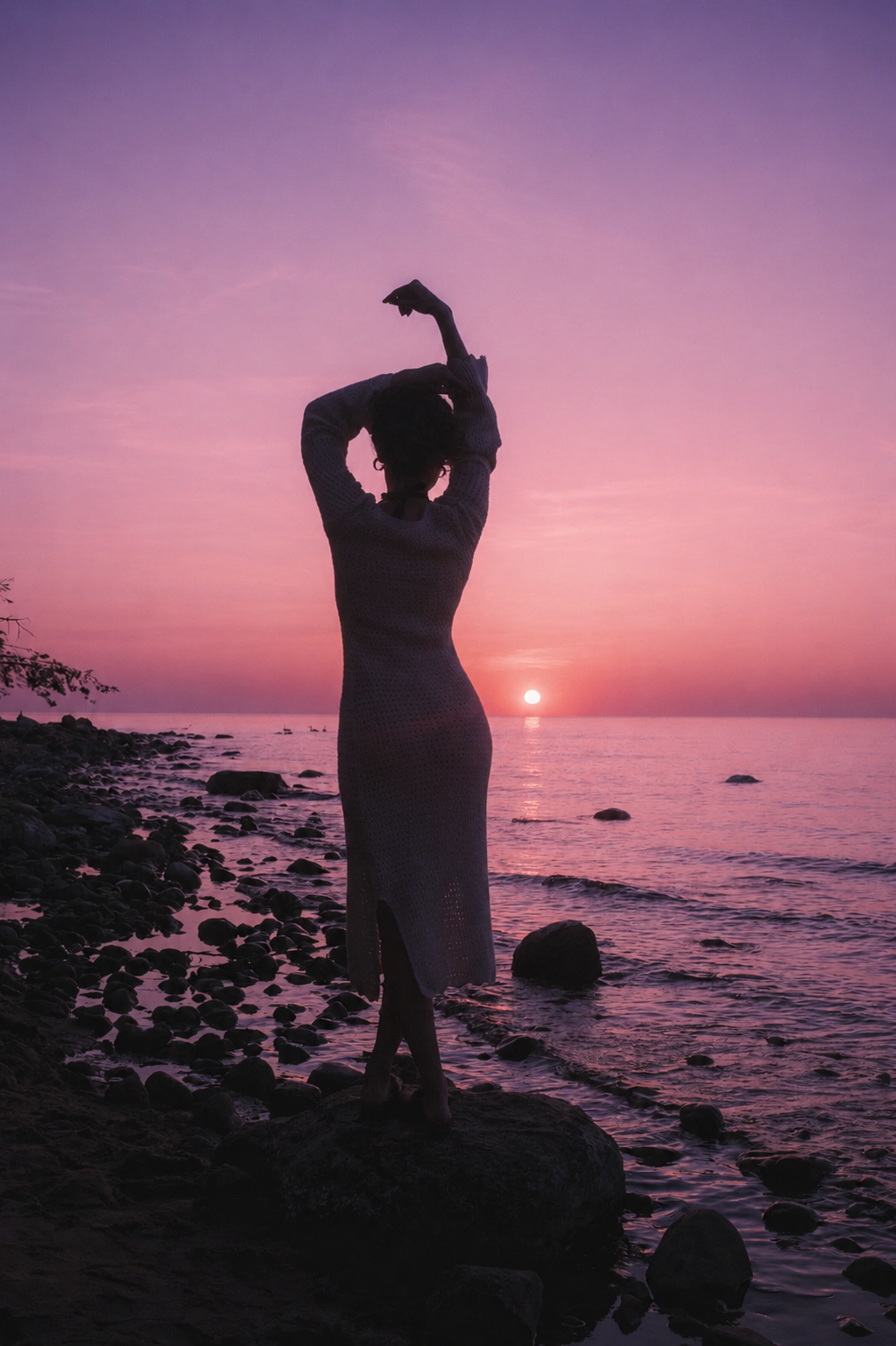 Woman standing by the water at sunset with her arms raised