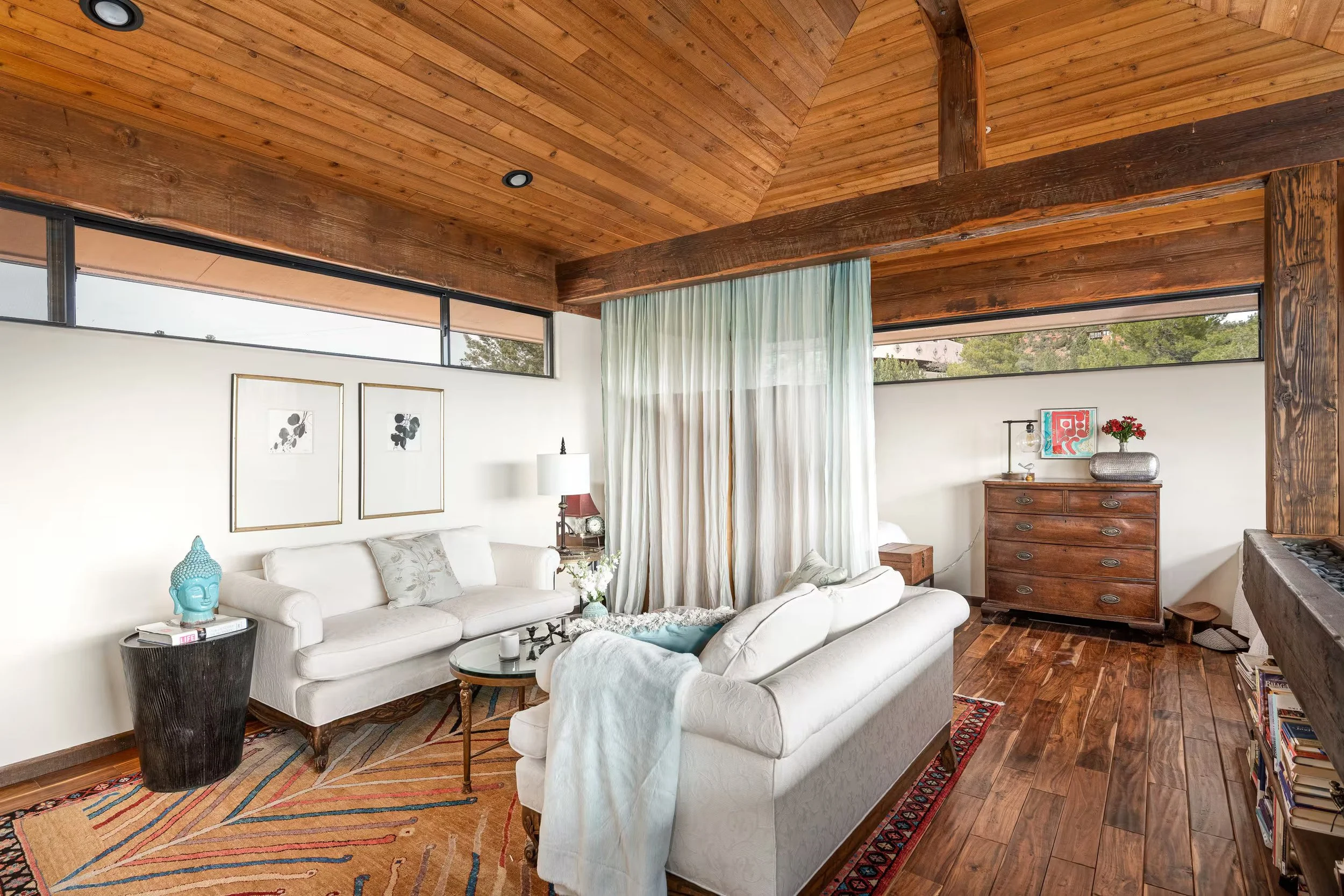 Living room with white sofas, wooden ceiling beams, framed black and white botanical prints, a side table with a turquoise Buddha head sculpture, and a large window with sheer curtains.