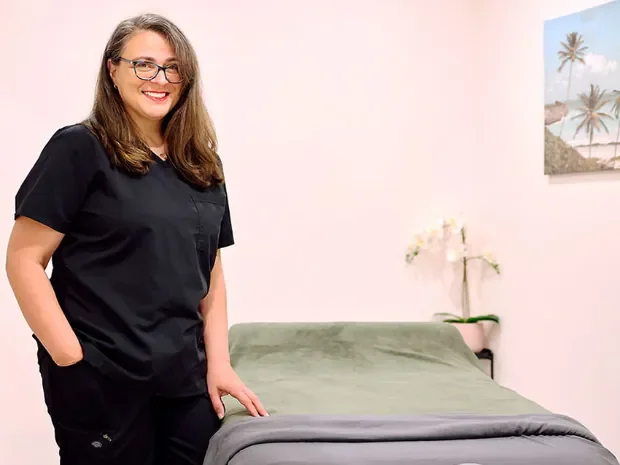 A woman with glasses and long brown hair smiling and wearing black scrubs, standing in a room with a bed and a picture of palm trees on the wall.