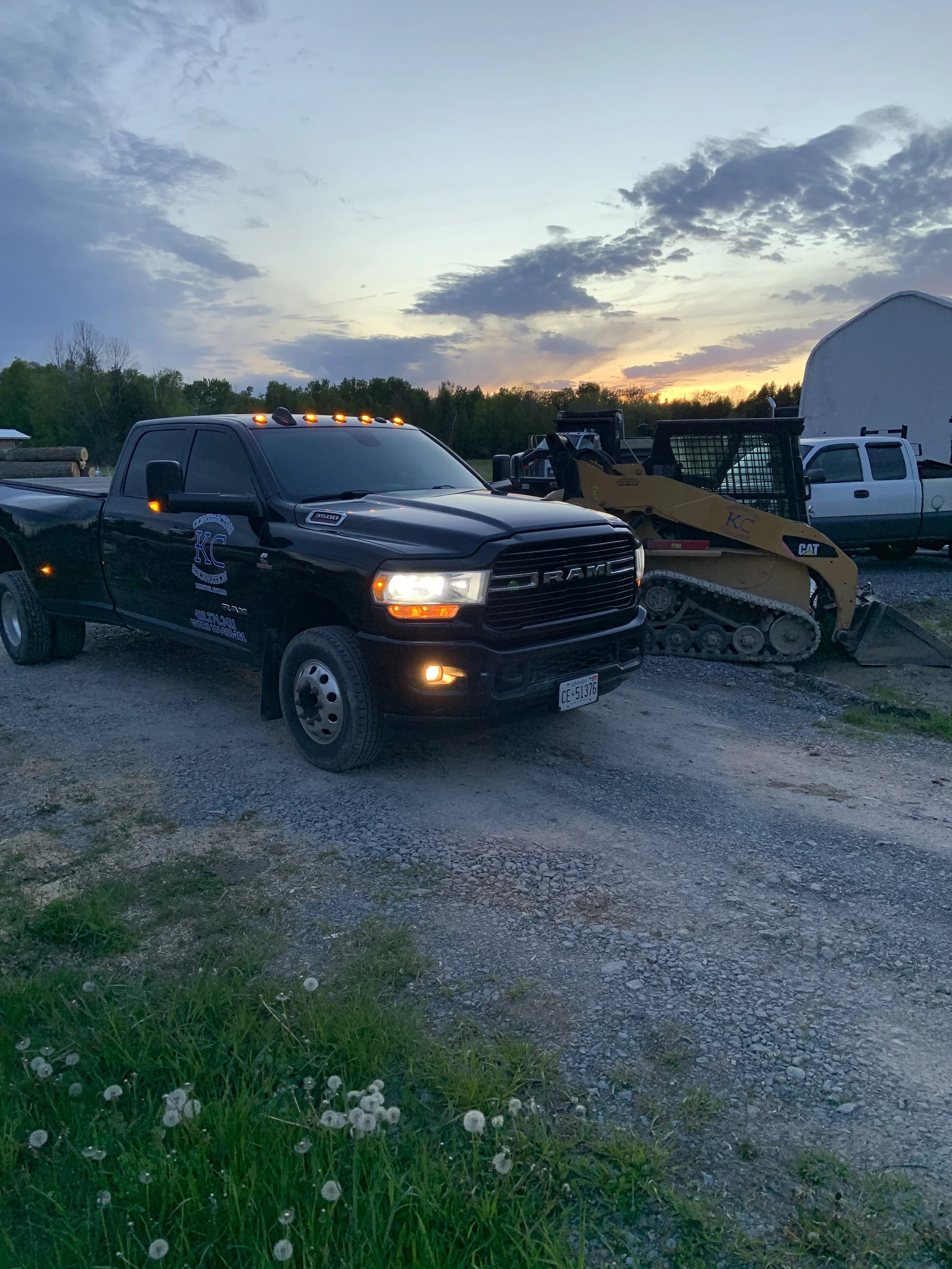A black RAM pickup truck with orange lights on the roof is parked on a gravel lot during sunset. Next to it is a small construction vehicle, and in the background, there is a white trailer and a white pickup truck. The sky is partly cloudy with a visible sunset near the horizon, and some grass and dandelions are visible in the foreground.
