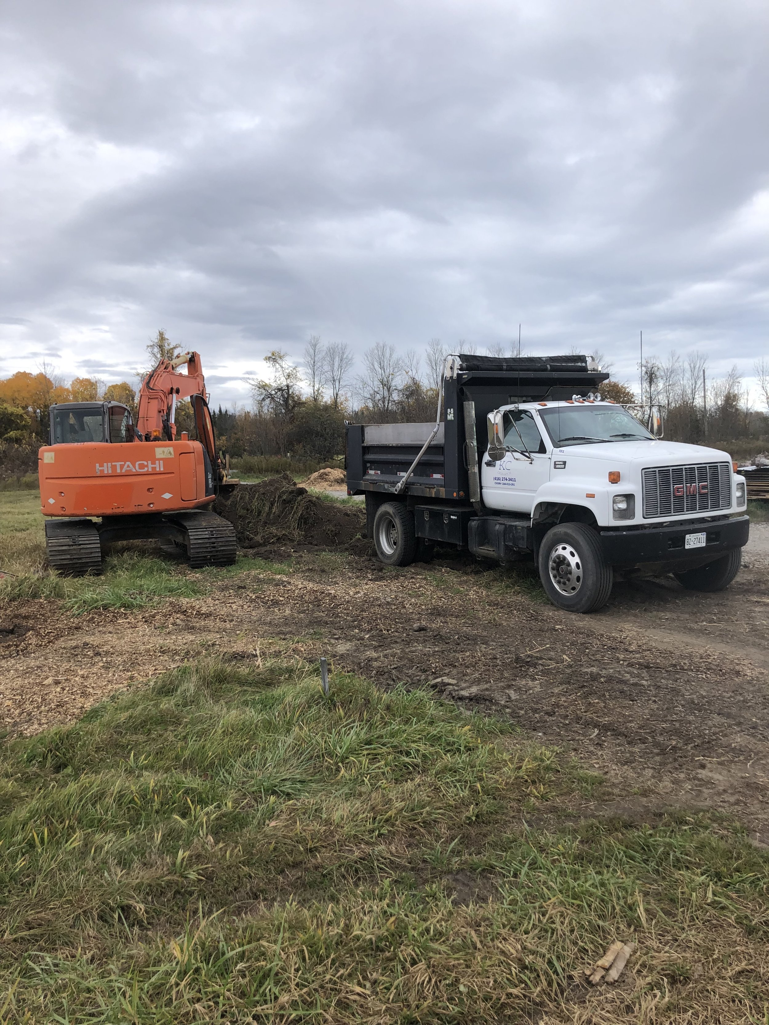A construction site with an orange Hitachi excavator digging into the ground and a white GMC dump truck parked nearby on muddy soil, with a background of trees and a cloudy sky.