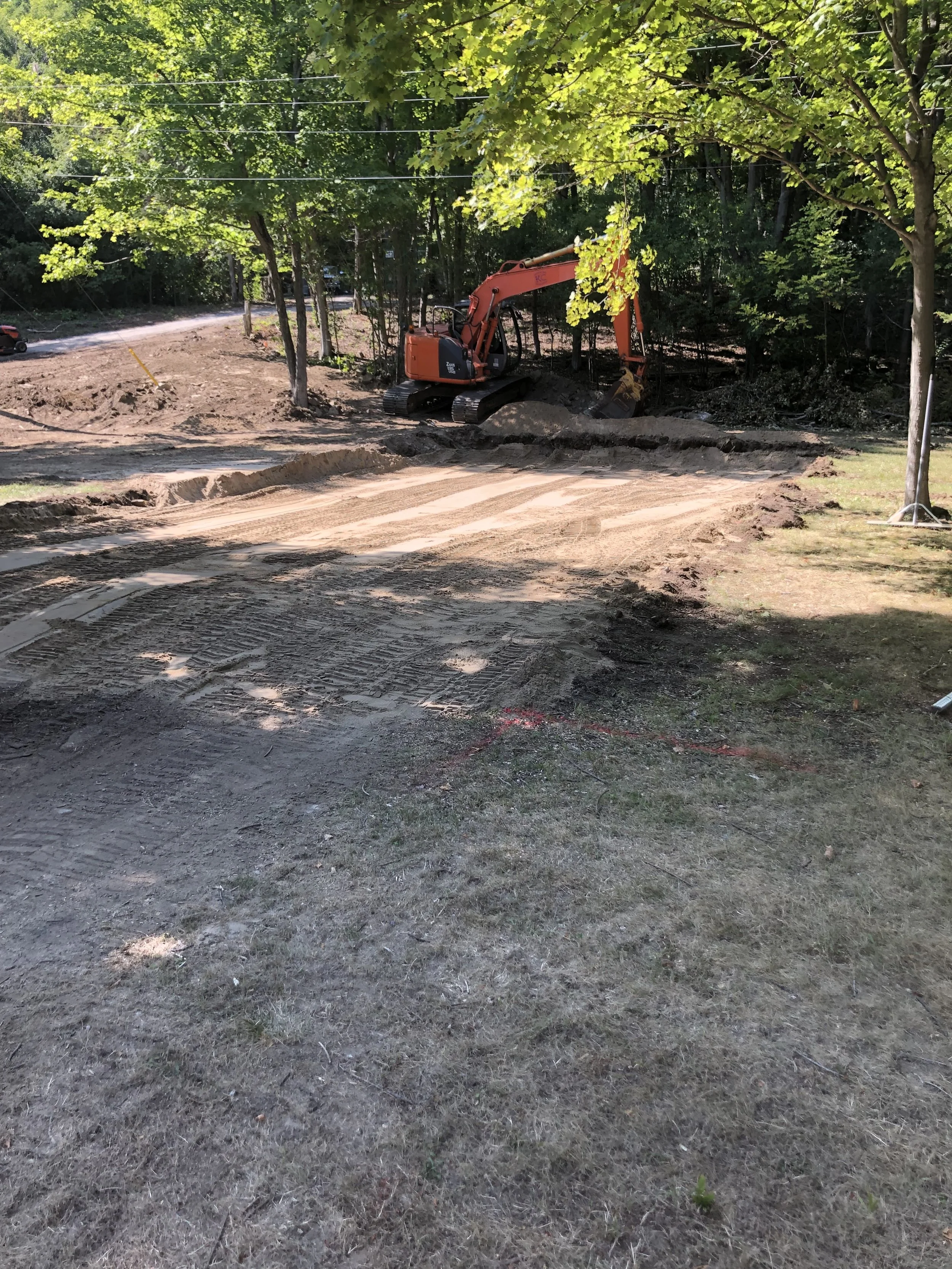 Construction site with dirt and tire tracks, orange excavator parked among trees, green leaves overhead, and a dirt road in the background.