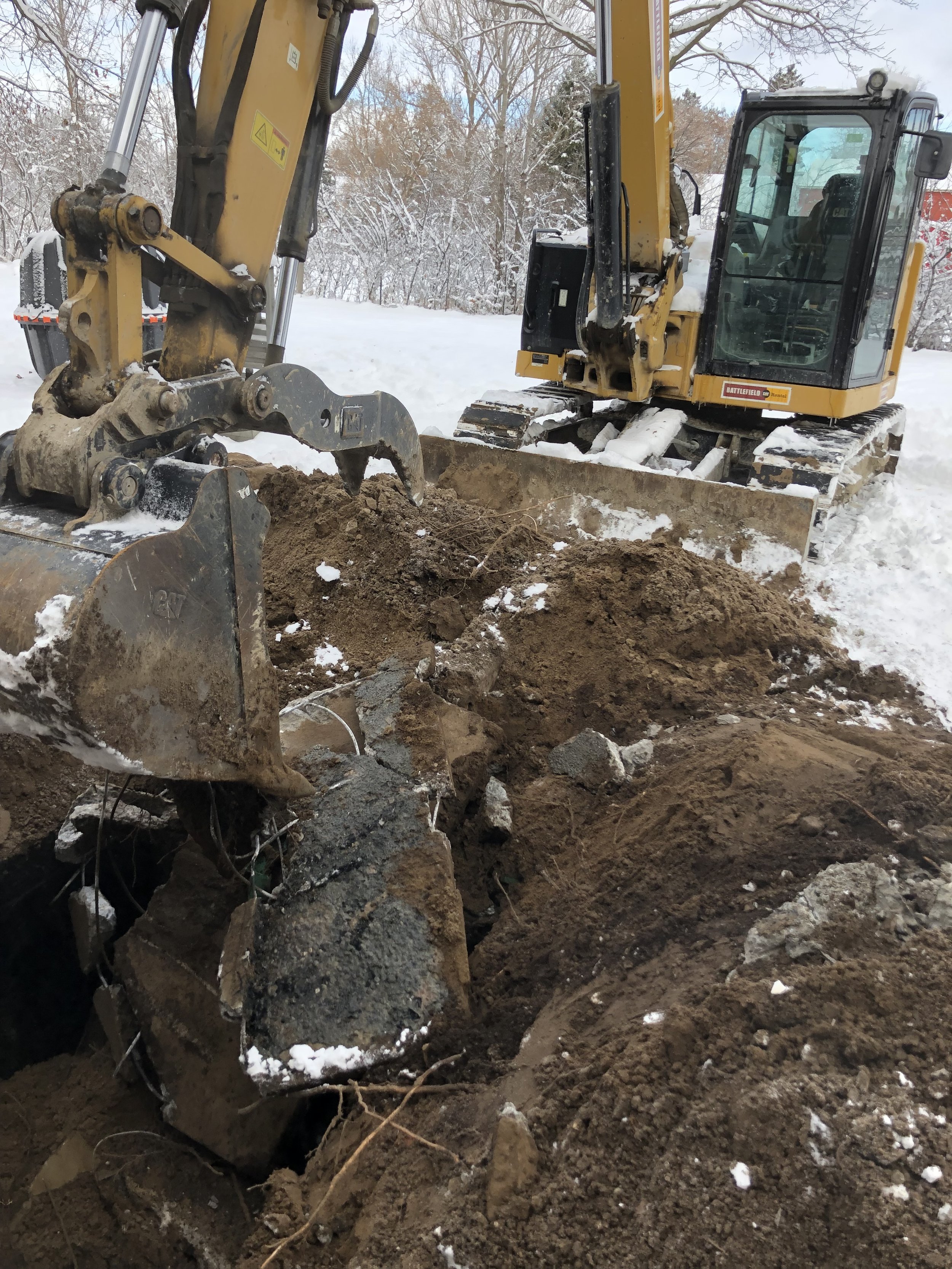 A small excavator working in a snowy outdoor environment, digging into the dirt and rocks.