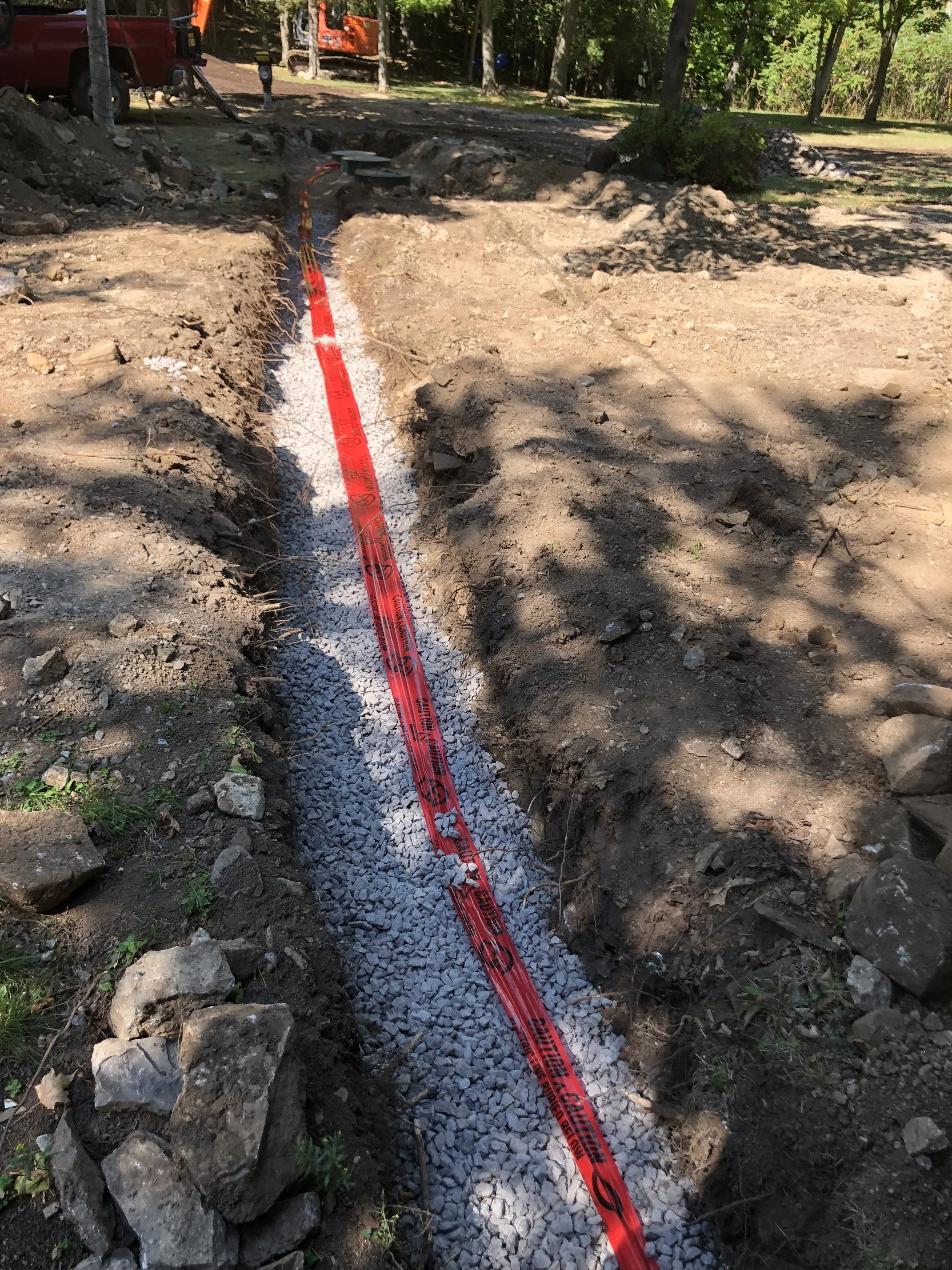 Construction site with a trench filled with gravel, orange and red safety tape, construction equipment, and trees in the background.