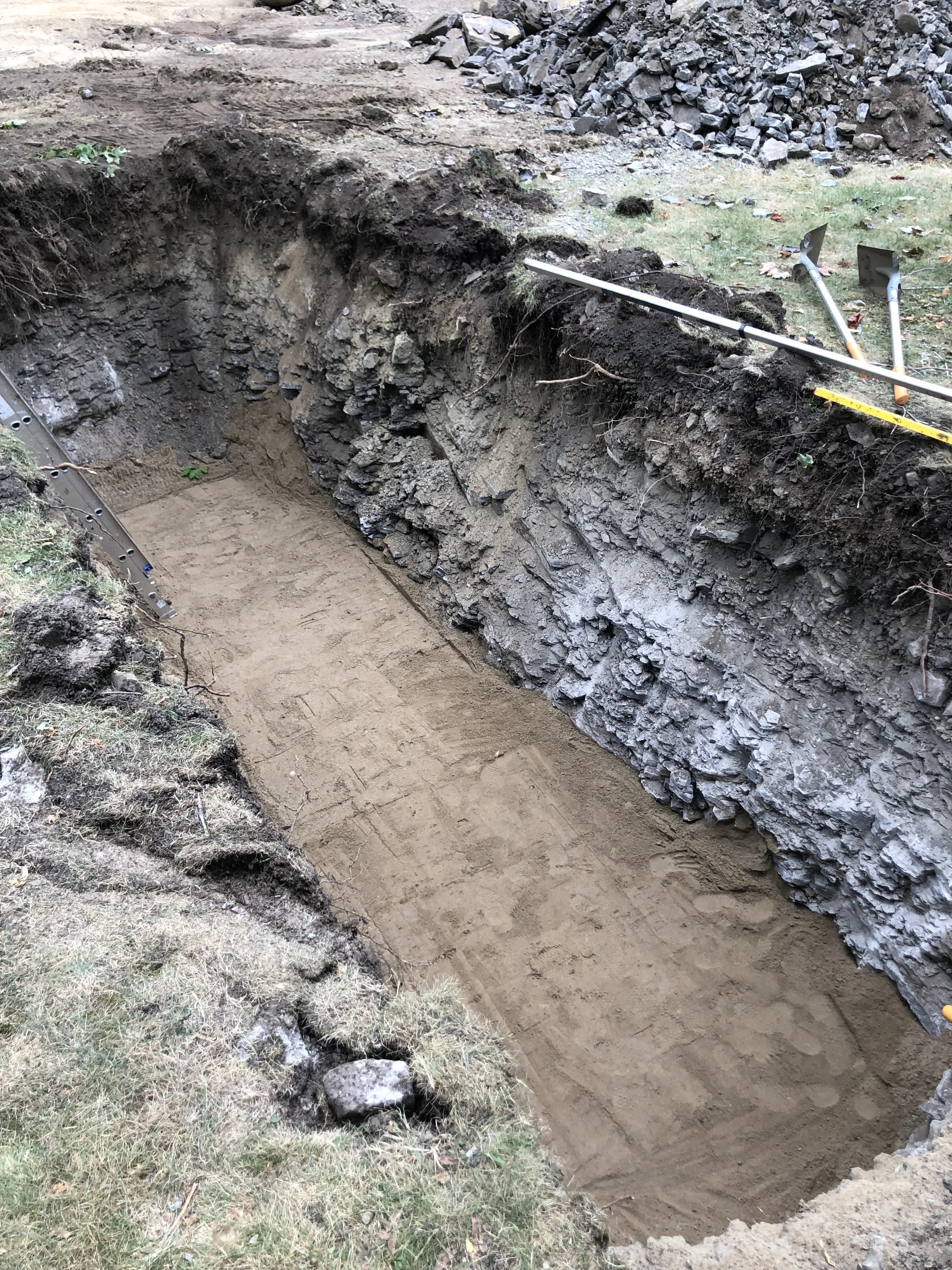 Excavation site showing a large trench with soil layers exposed on the side, along with construction tools like shovels and a level, and some scattered rocks and grass around the site.
