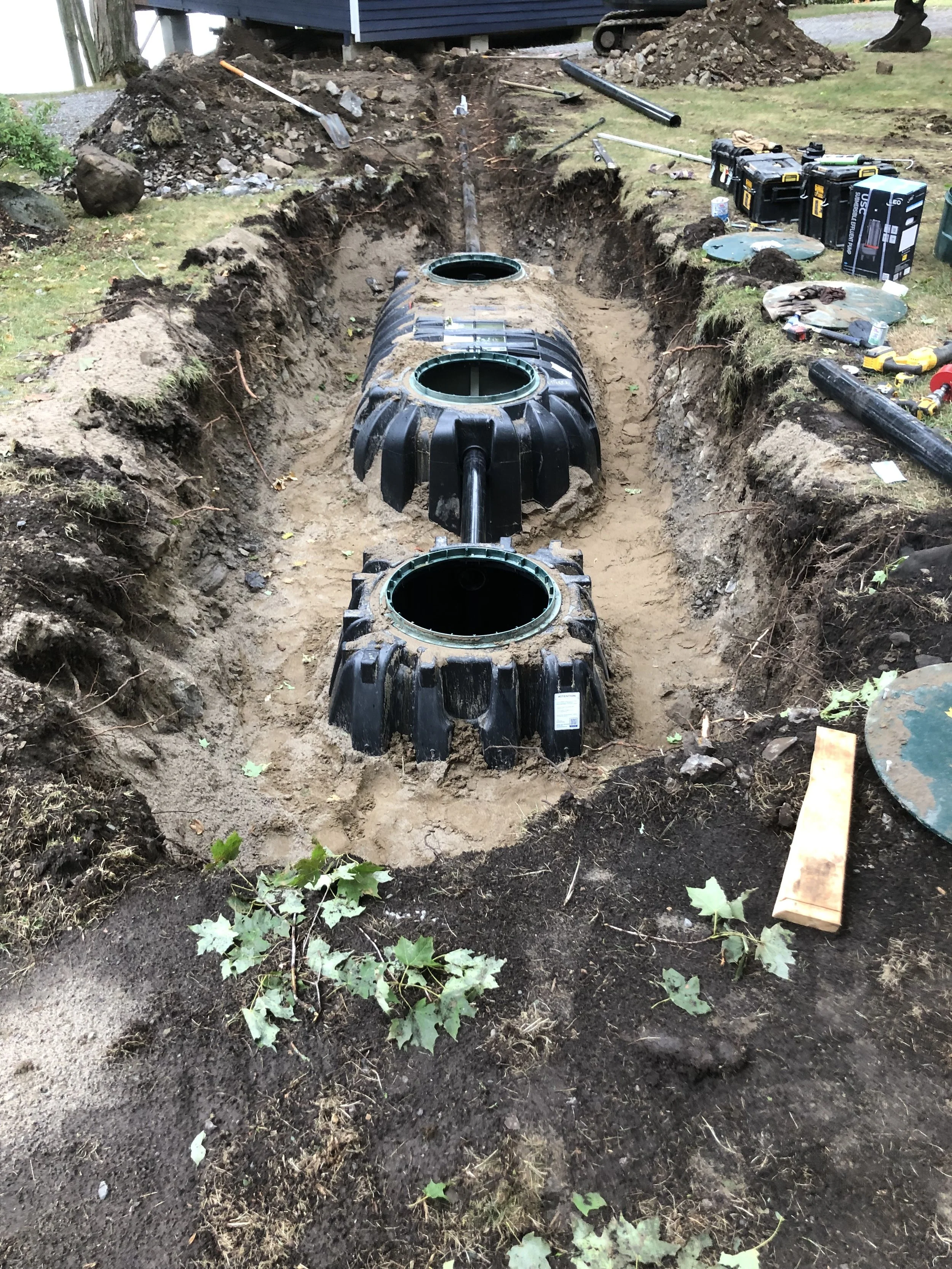 Dig trench for underground piping or drainage system with large black plastic pipes in a yard, construction tools and equipment scattered around.