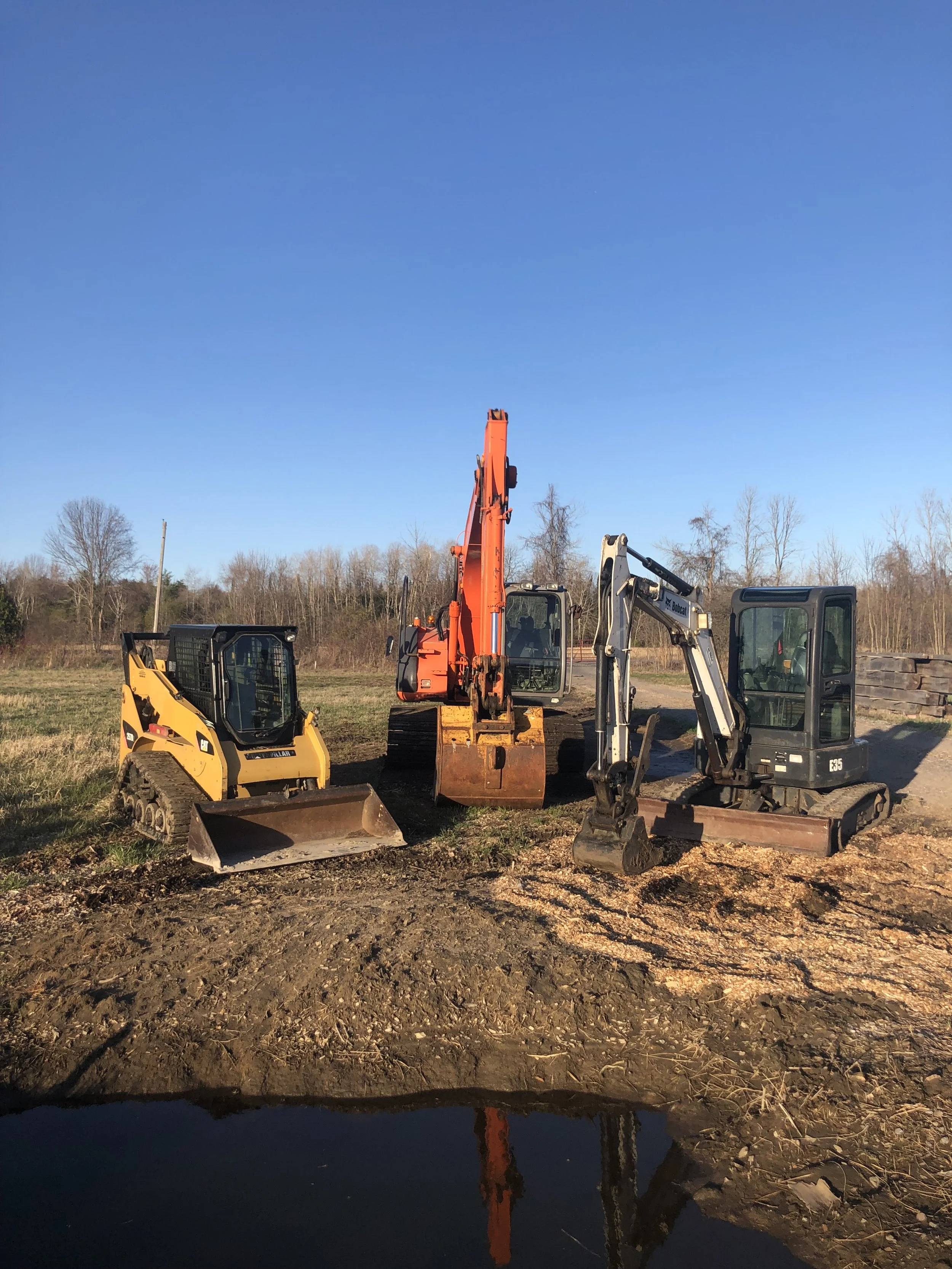 Construction site with multiple excavators and earth-moving equipment on a dirt field, with a clear blue sky and leafless trees in the background.