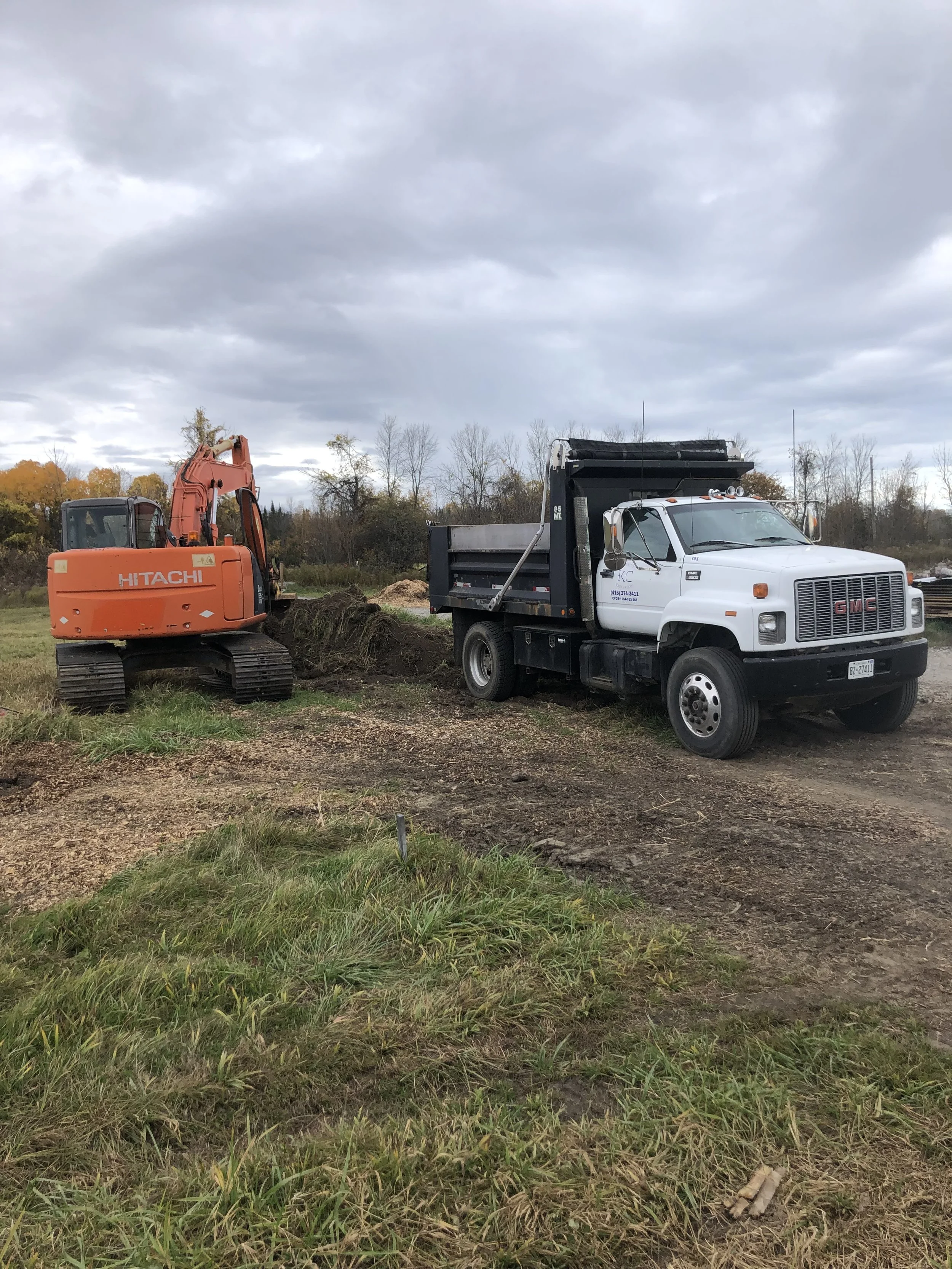 A construction site with an orange Hitachi excavator digging in the dirt and a white GMC dump truck parked nearby. Trees with autumn foliage and a cloudy sky in the background.