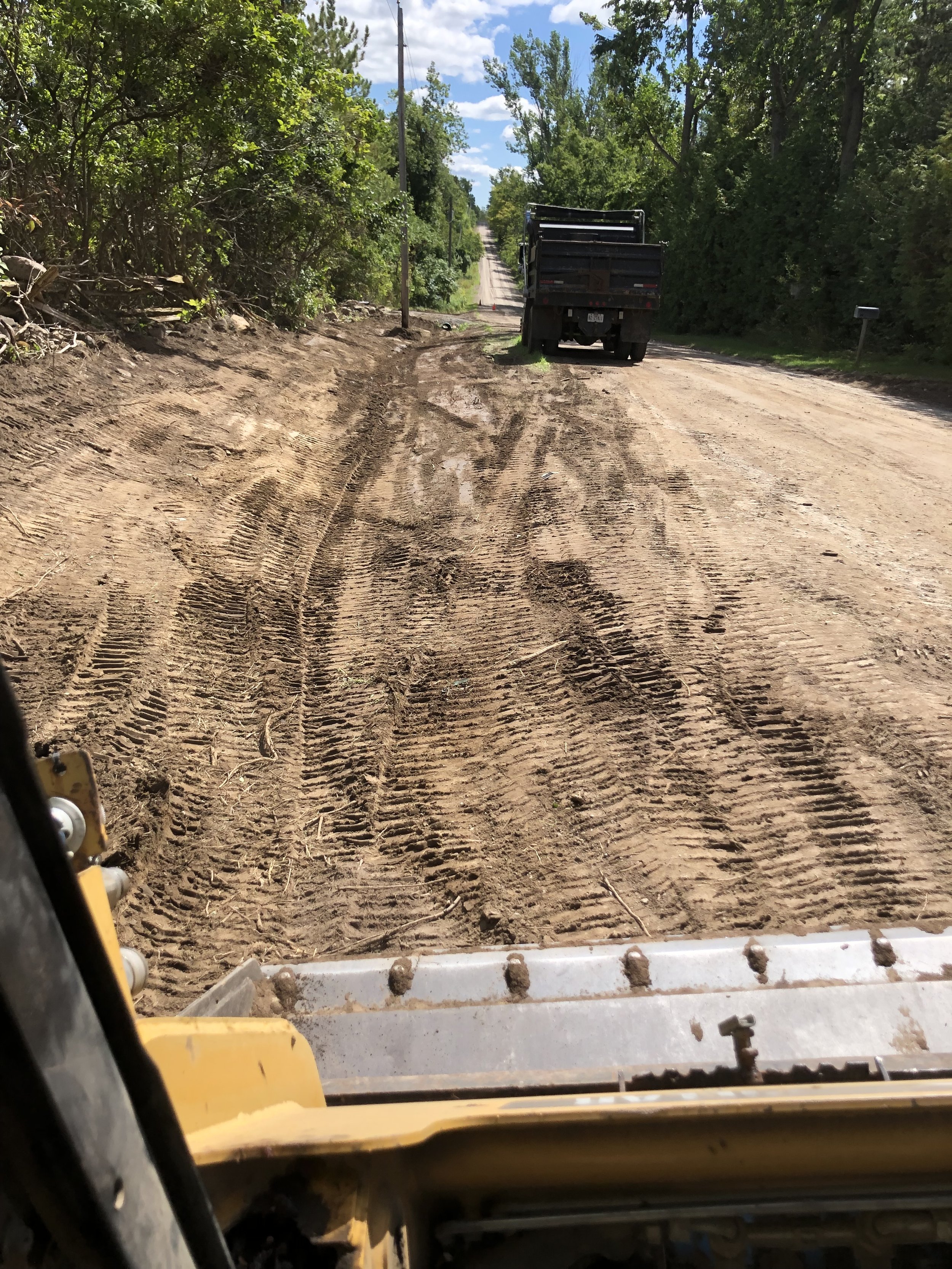 View from inside a construction vehicle showing a dirt road under repair with tire tracks, green trees on either side, and a partly cloudy sky overhead.