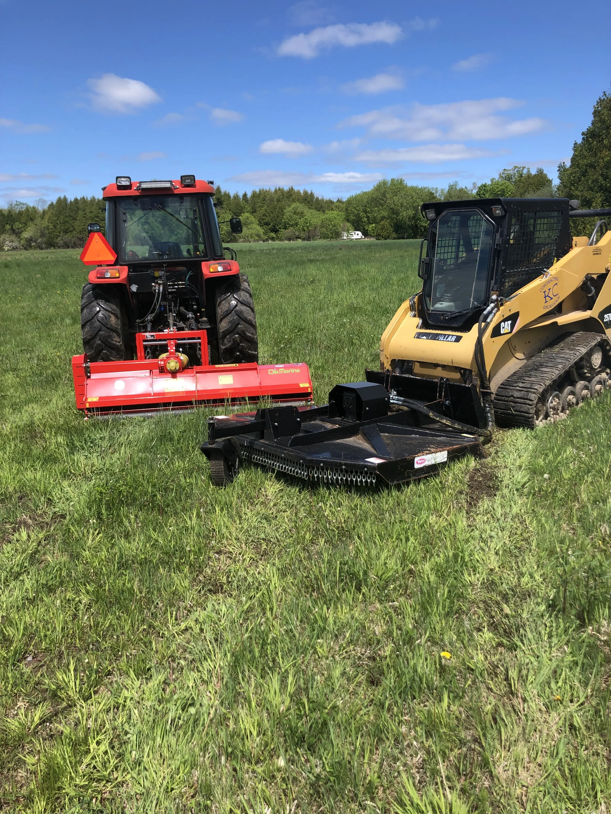 A red tractor and a yellow skid steer loader working on a grassy field under a blue sky with clouds, possibly for land clearing, brush hogging, or mowing