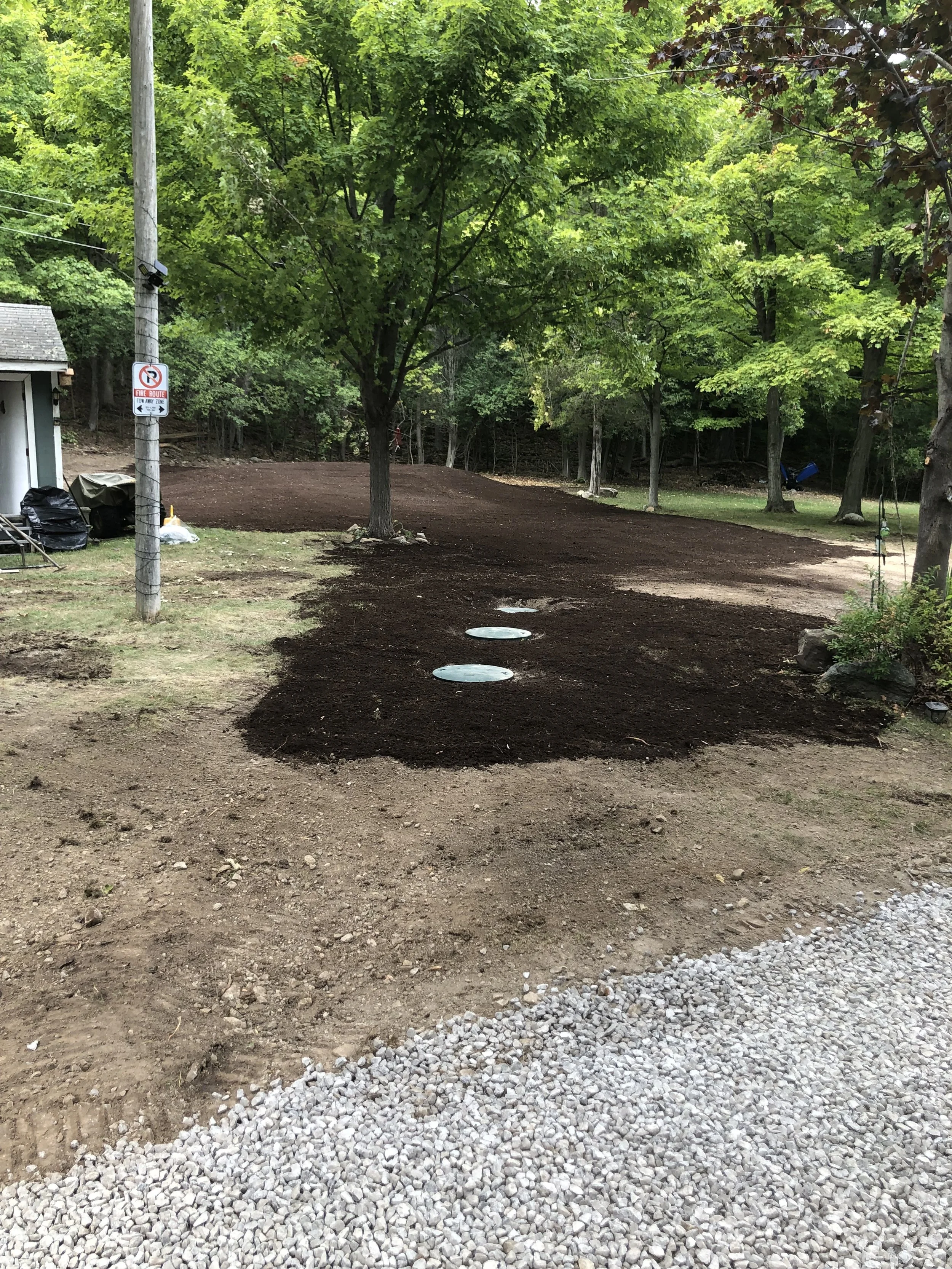 A backyard with trees, a utility pole, and a partially landscaped area with dark soil, gravel, and stepping stones.