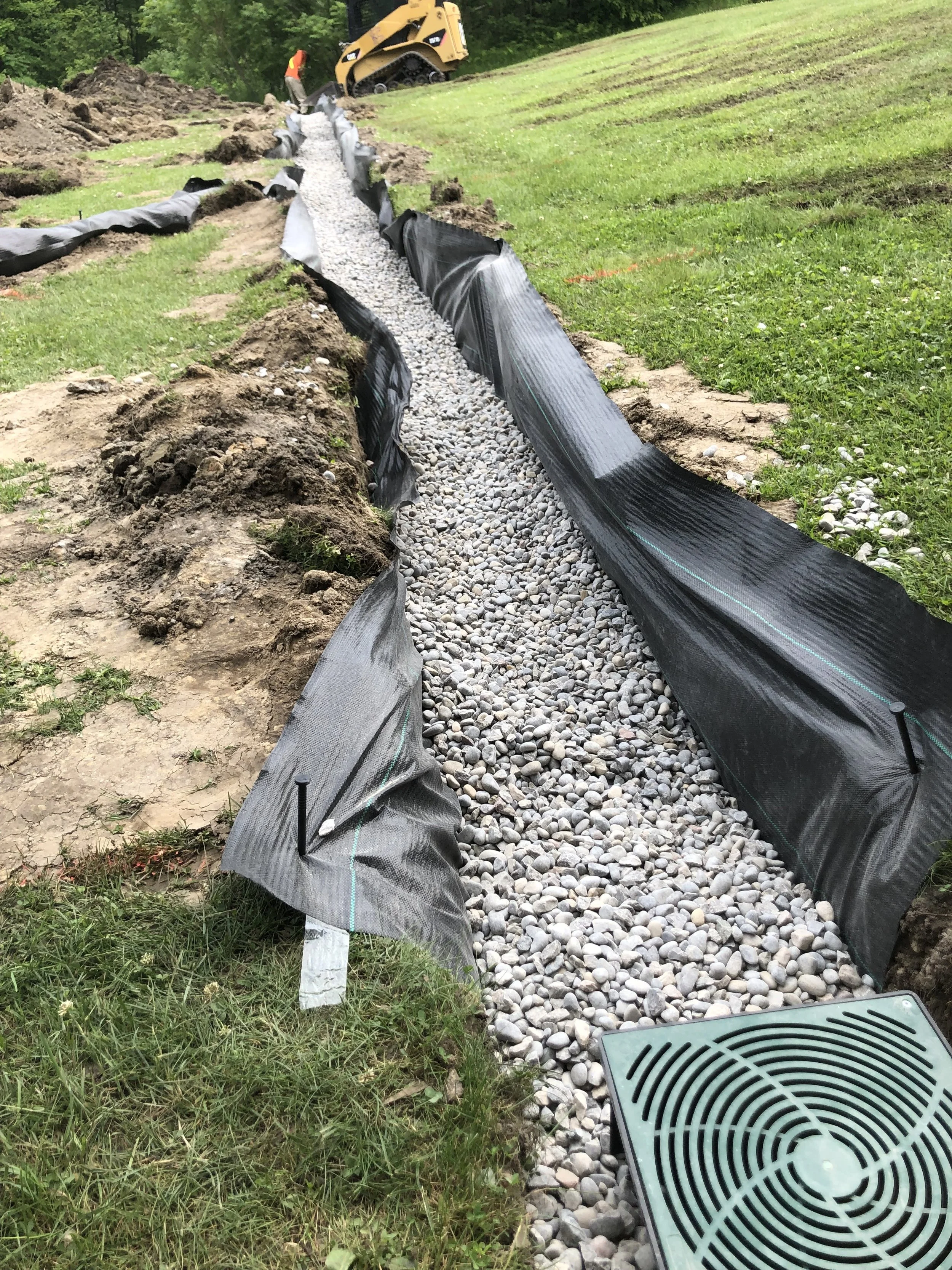 Construction site with a gravel drainage system being installed along a grassy slope, black landscape fabric lining the trench, a mini bulldozer, and a drainage grate in the foreground.
