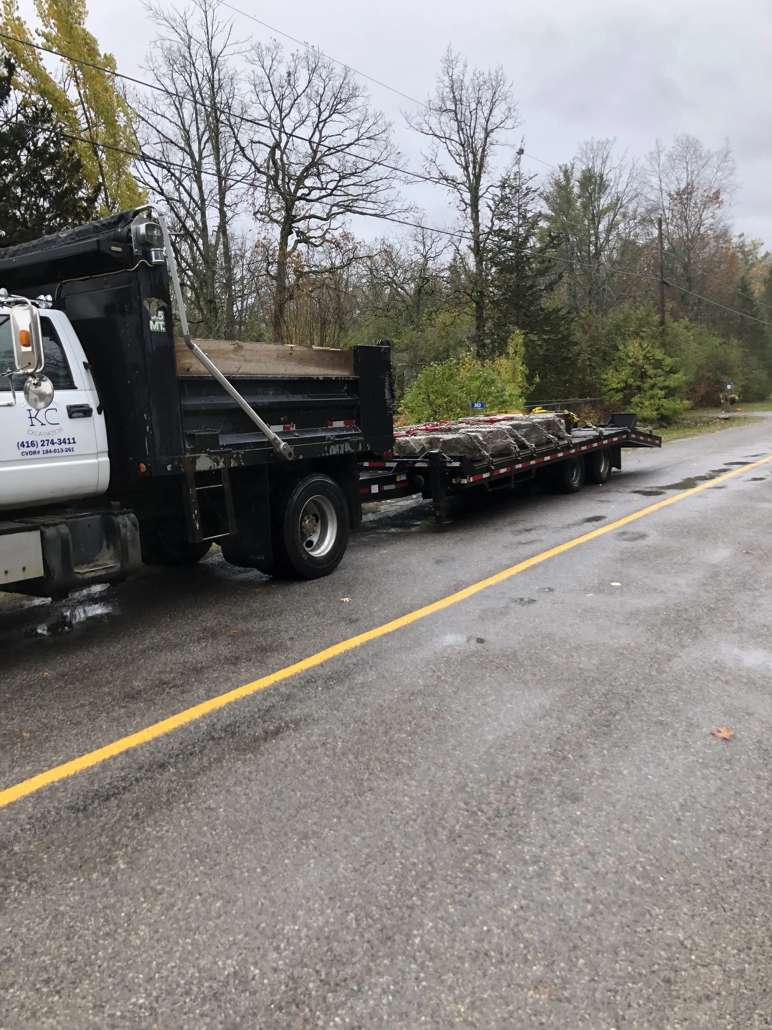 A flatbed truck parked on a wet road with bare trees and overcast sky in the background.
