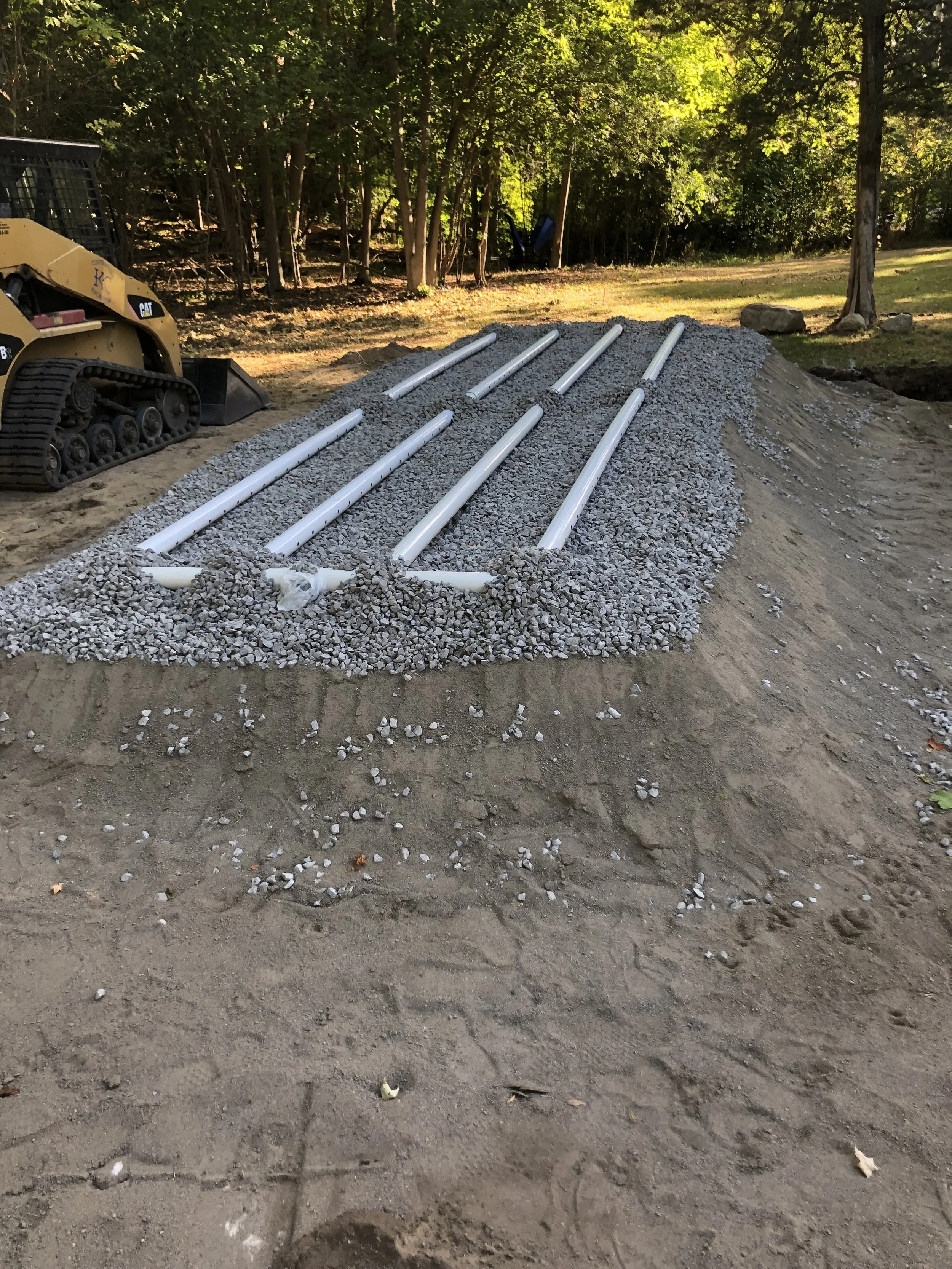 Construction site with a small bulldozer on the left and white pipes laid on a bed of gravel, preparing for a pathway or foundation, with trees and a green wooded area in the background.