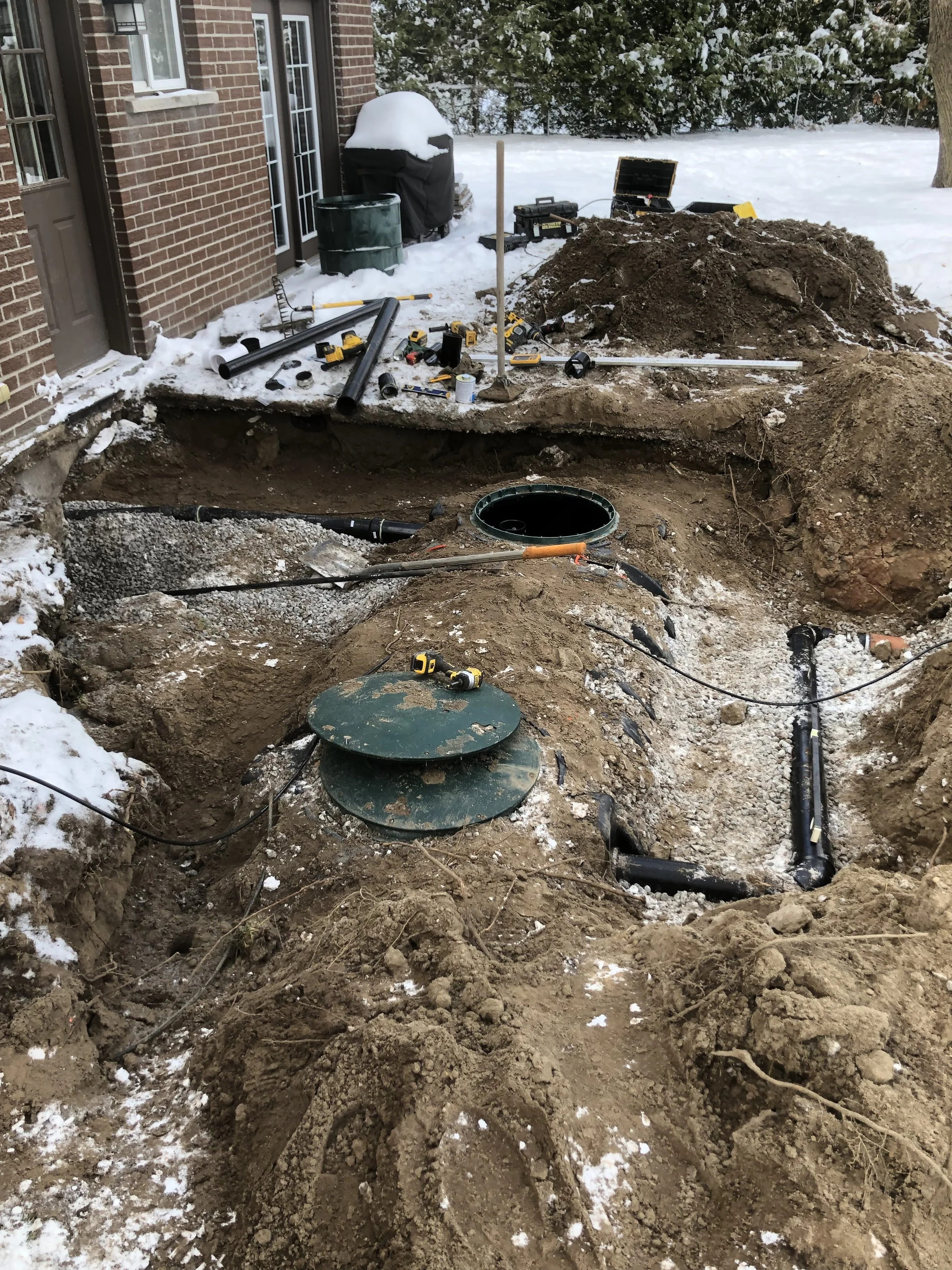 Underground piping installation in a residential yard, with tools, soil, and a hole exposing pipes near a brick house, snow on the ground.